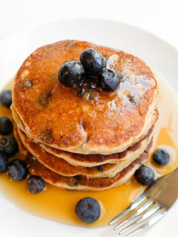 A stack of gluten-free pancakes on a white plate. Blueberries and maple syrup are topping the pancakes.