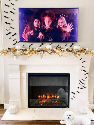 A fireplace decorated for Halloween. Black bat stickers are flying up the fireplace and the wall. A tv is hung above the fireplace and a cute white dog names CoCo lays in front of the fireplace.