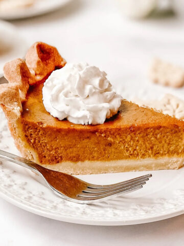 A photo showing a slice of my gluten-free pumpkin pie. The slice is placed on a white decorative plate with a silver fork. There is homemade whipped cream on top of the pie.