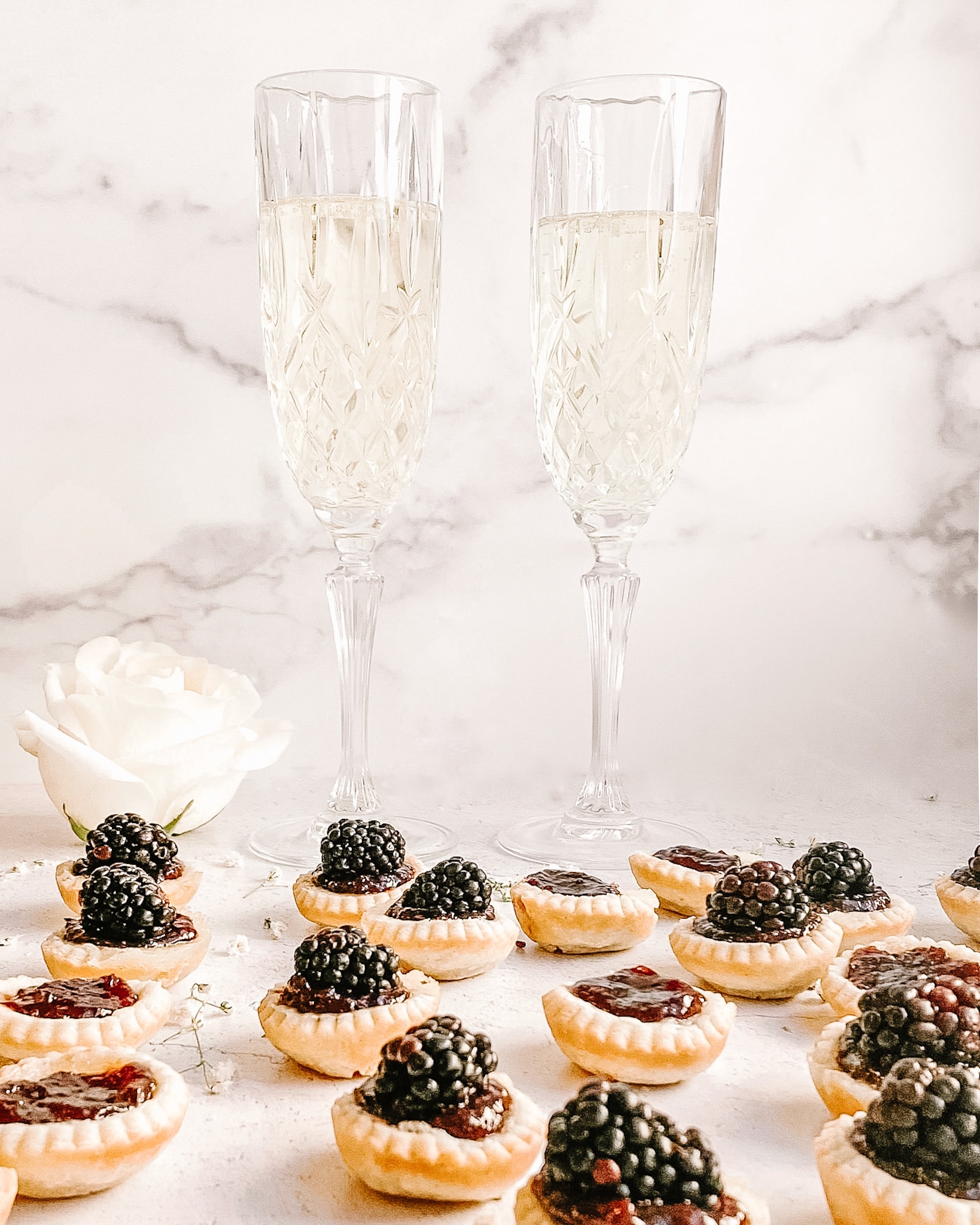 a spread of mini blackberry tarts, two champagne glasses with champagne in the background