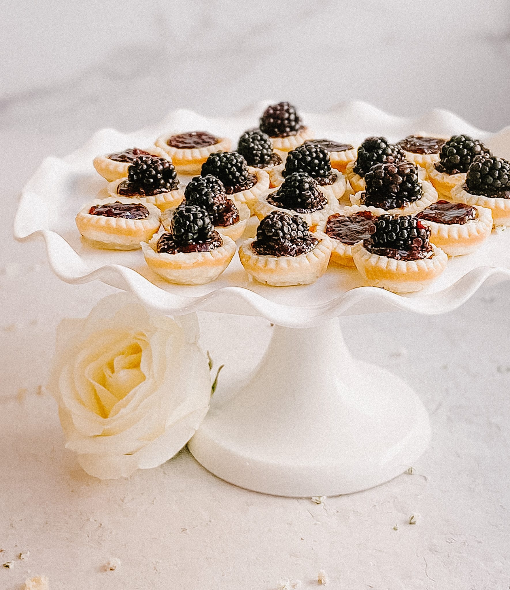 gluten-free blackberry tarts on a white cake stand, white flowers adjourn the base of the stand