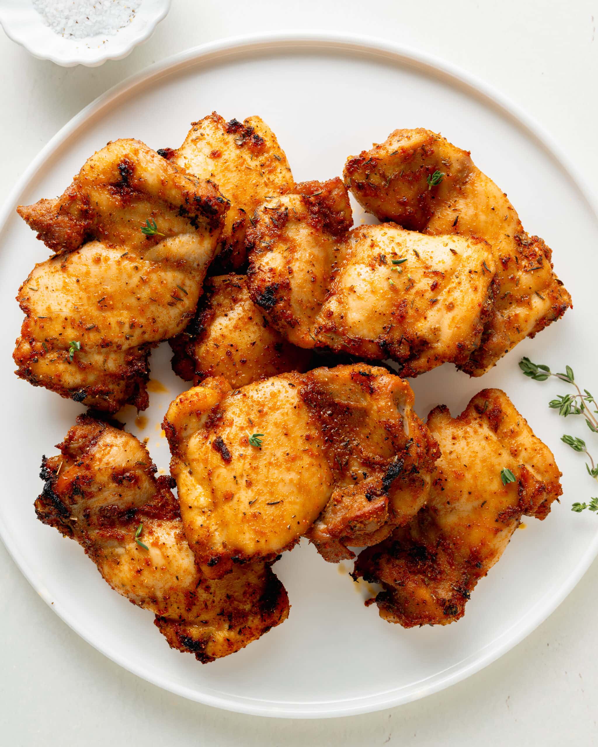 A picture of boneless skinless chicken thighs on a white plate with a small white bowl of salt and pepper, and fresh thyme sprinkled on top of the chicken.