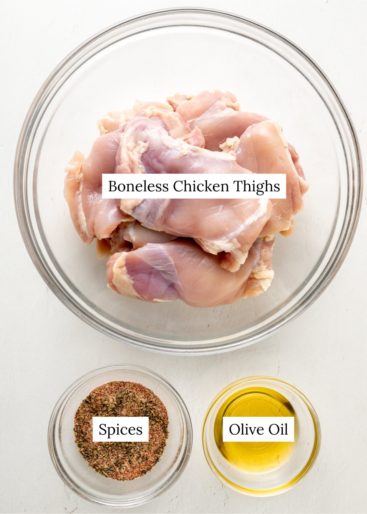 Boneless skinless chicken thighs in a large clear bowl, next to a smaller bowl of olive oil and another bowl full of spices.