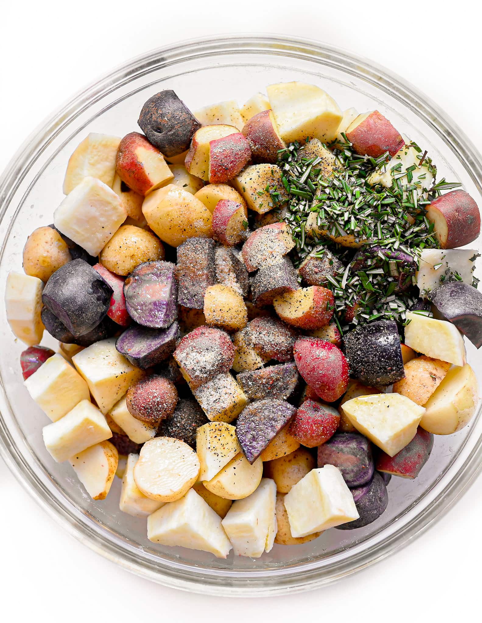 Bowl of uncooked mixed colored potatoes. Fresh seasoning and rosemary is on the potatoes.
