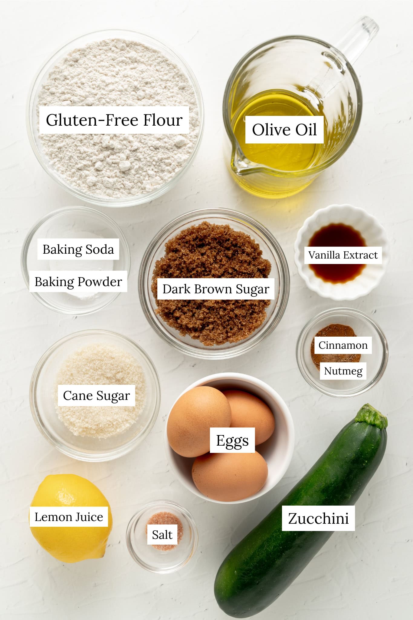 Overhead view of ingredients for gluten-free zucchini bread, including a fresh whole zucchini, three brown eggs, dark brown sugar, gluten-free flour, olive oil, granulated sugar, a lemon, vanilla extract, and spices arranged in small glass bowls on a white background.