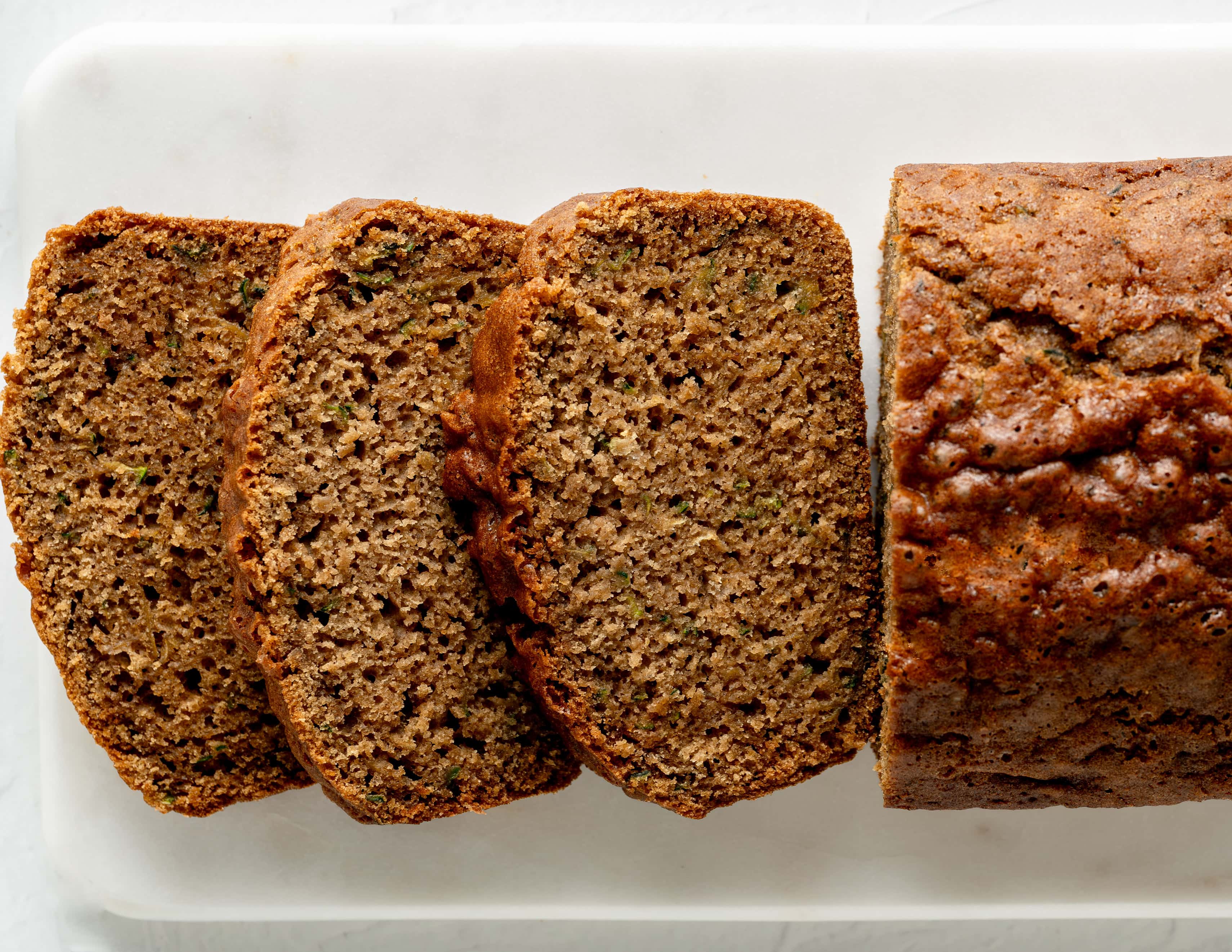 Overhead view of a loaf of gluten-free zucchini bread sliced on a white marble cutting board, showing a moist, tender crumb with golden-brown crust.