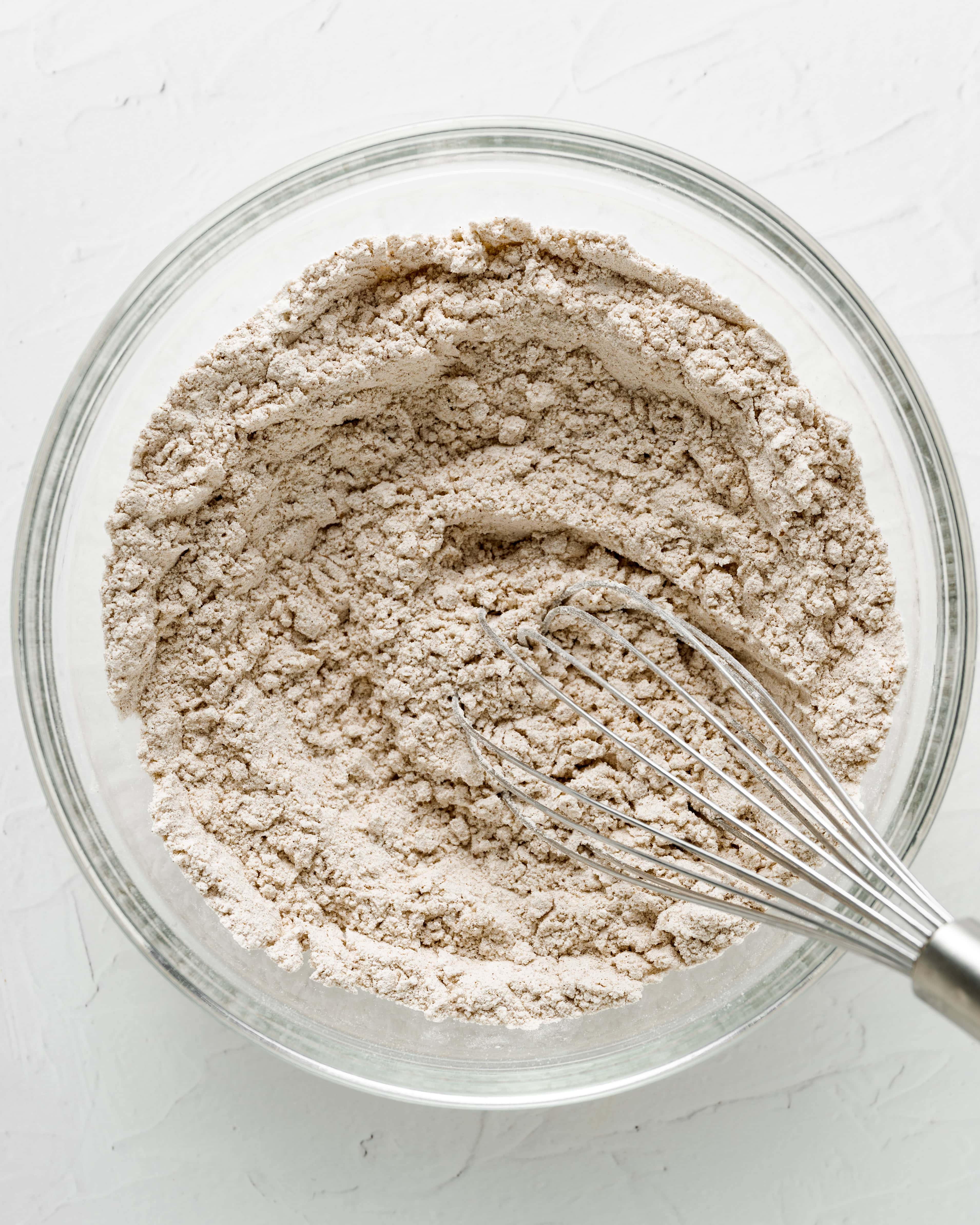 Overhead view of a wire whisk mixing gluten-free flour, warm spices, baking soda, and salt together in a clear glass bowl for Step 1 of making zucchini bread.