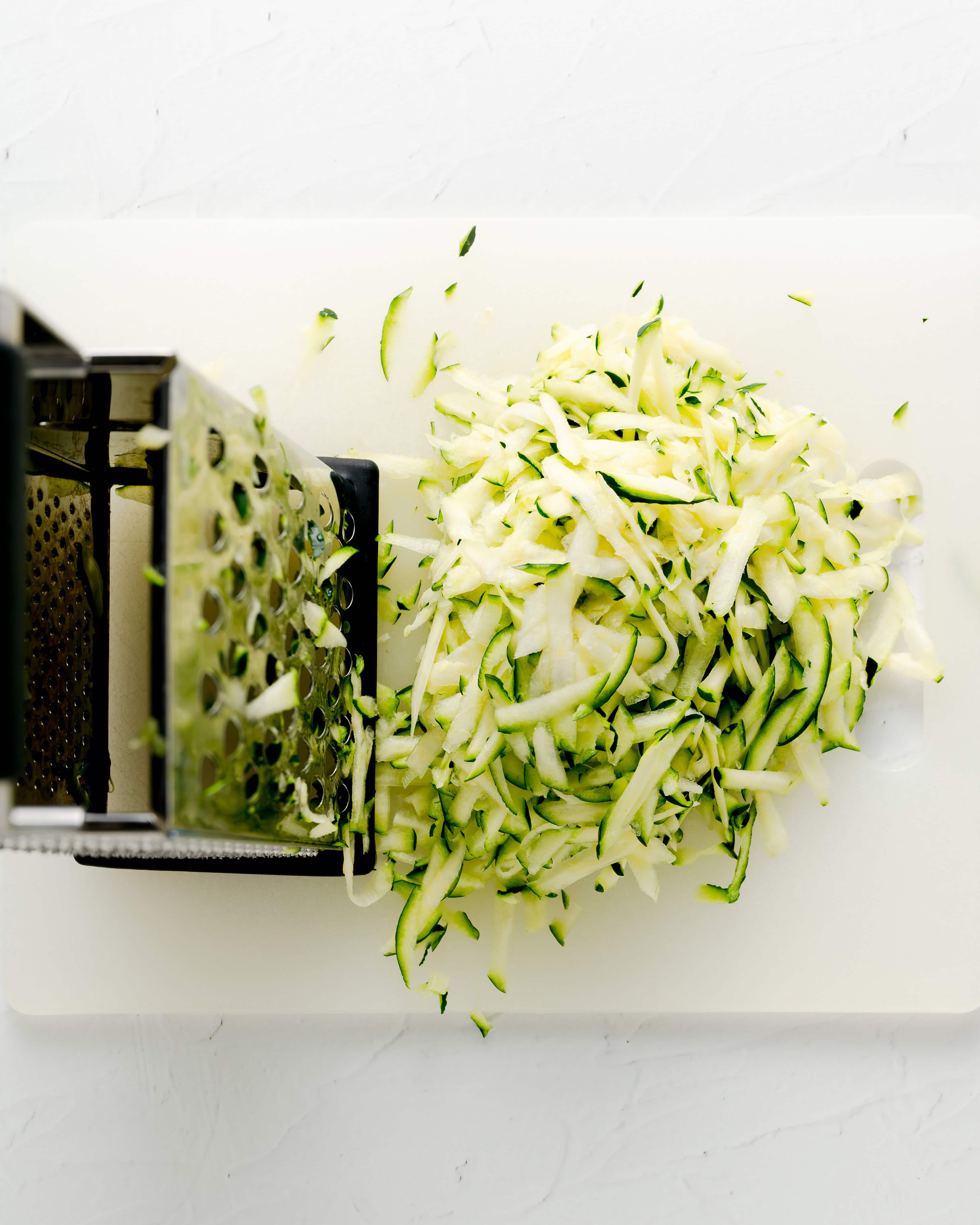 overhead view of freshly shredded green zucchini piled on a white cutting board next to a stainless steel box grater.