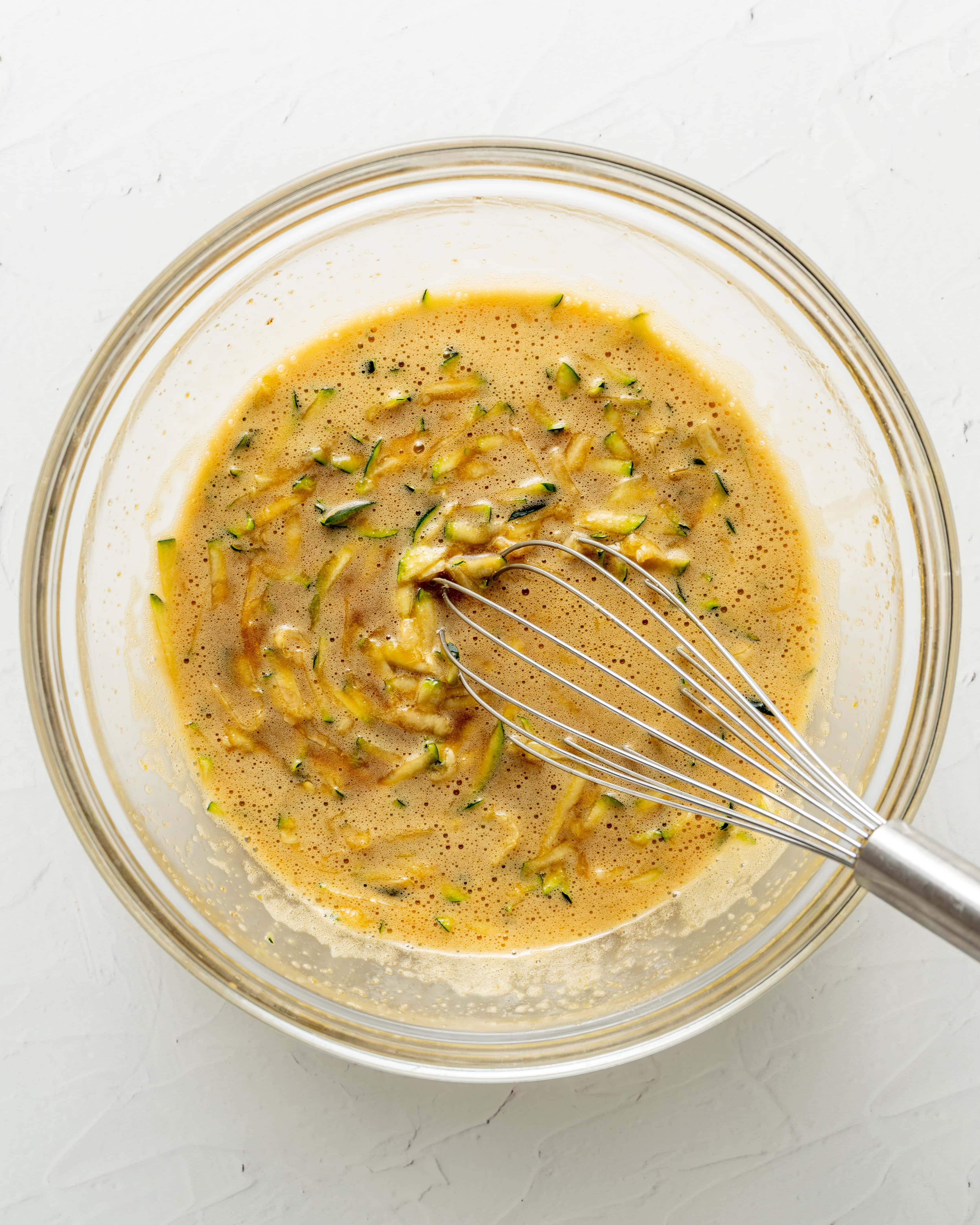 Overhead view of a wire whisk combining grated zucchini with the wet ingredients in a clear glass mixing bowl for Step 3 of making zucchini bread.