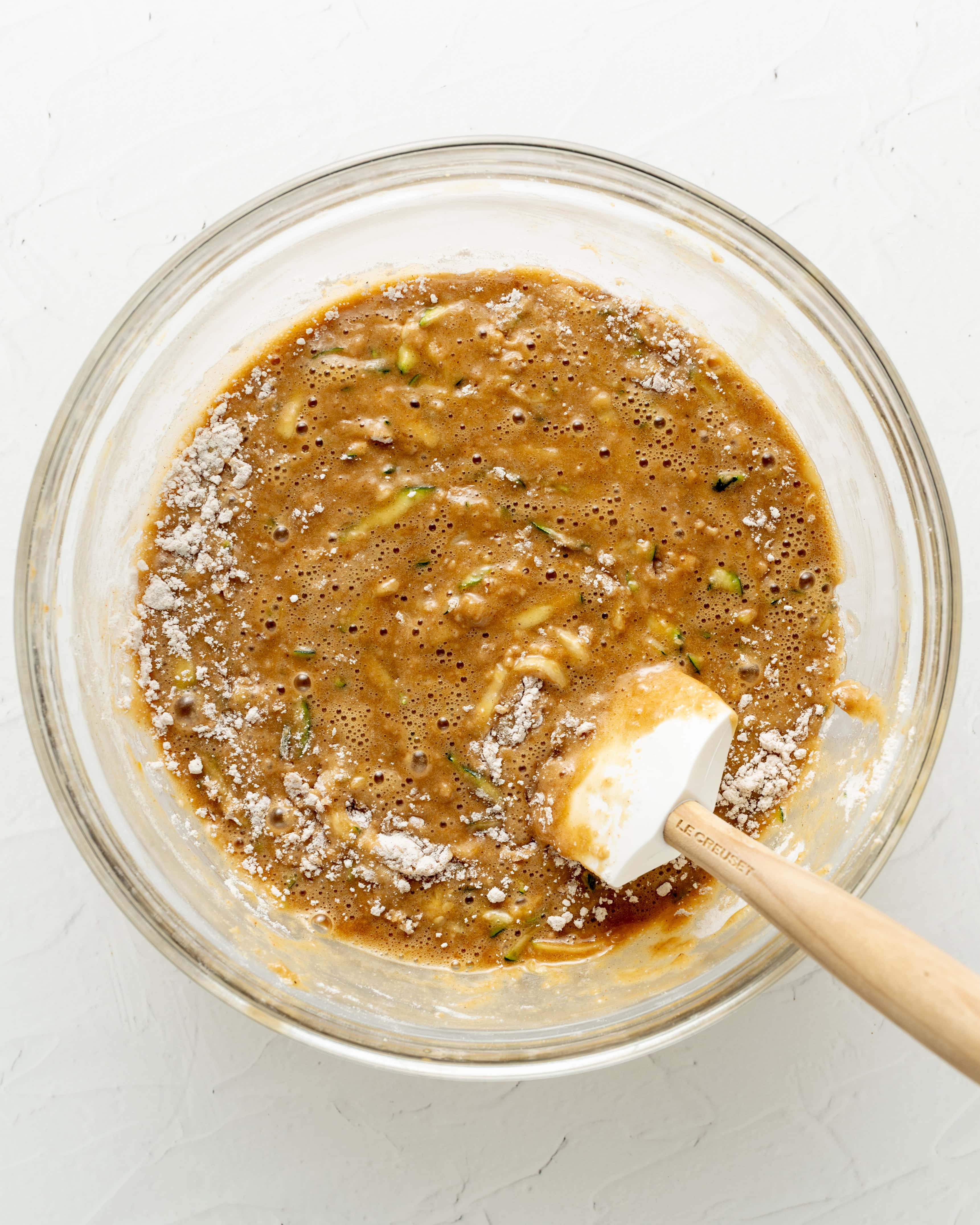 Overhead view of a white silicone spatula folding wet and dry ingredients together in a glass bowl to form gluten-free zucchini bread batter.