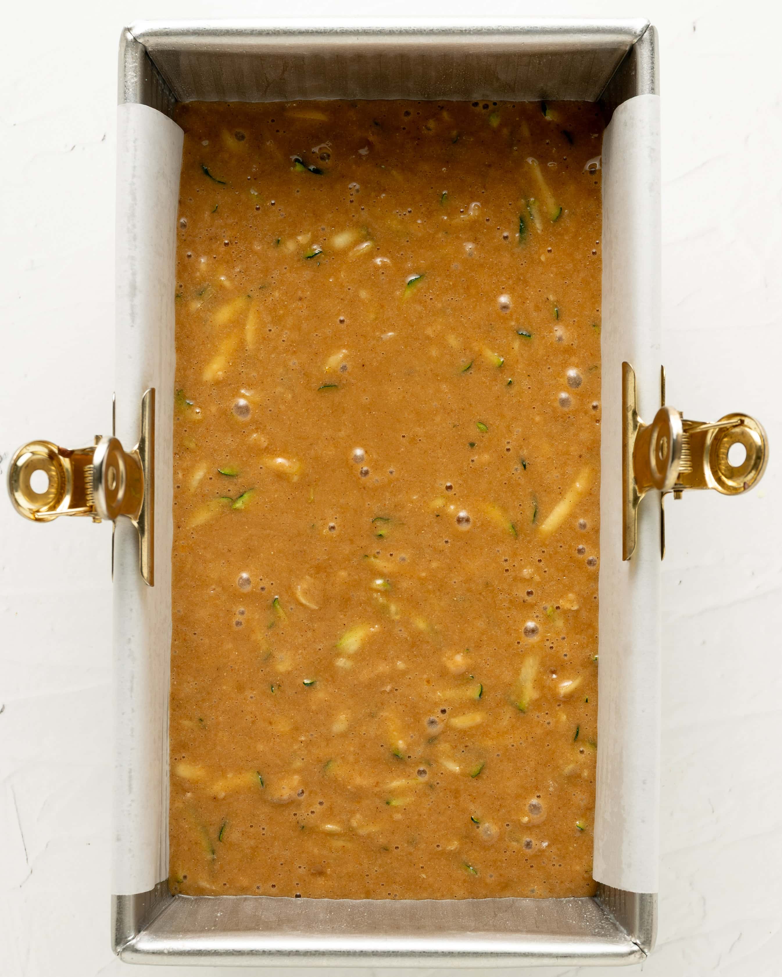 Overhead view of raw gluten-free zucchini bread batter poured into a parchment-lined metal loaf pan secured with gold binder clips, resting for 20 minutes before baking.