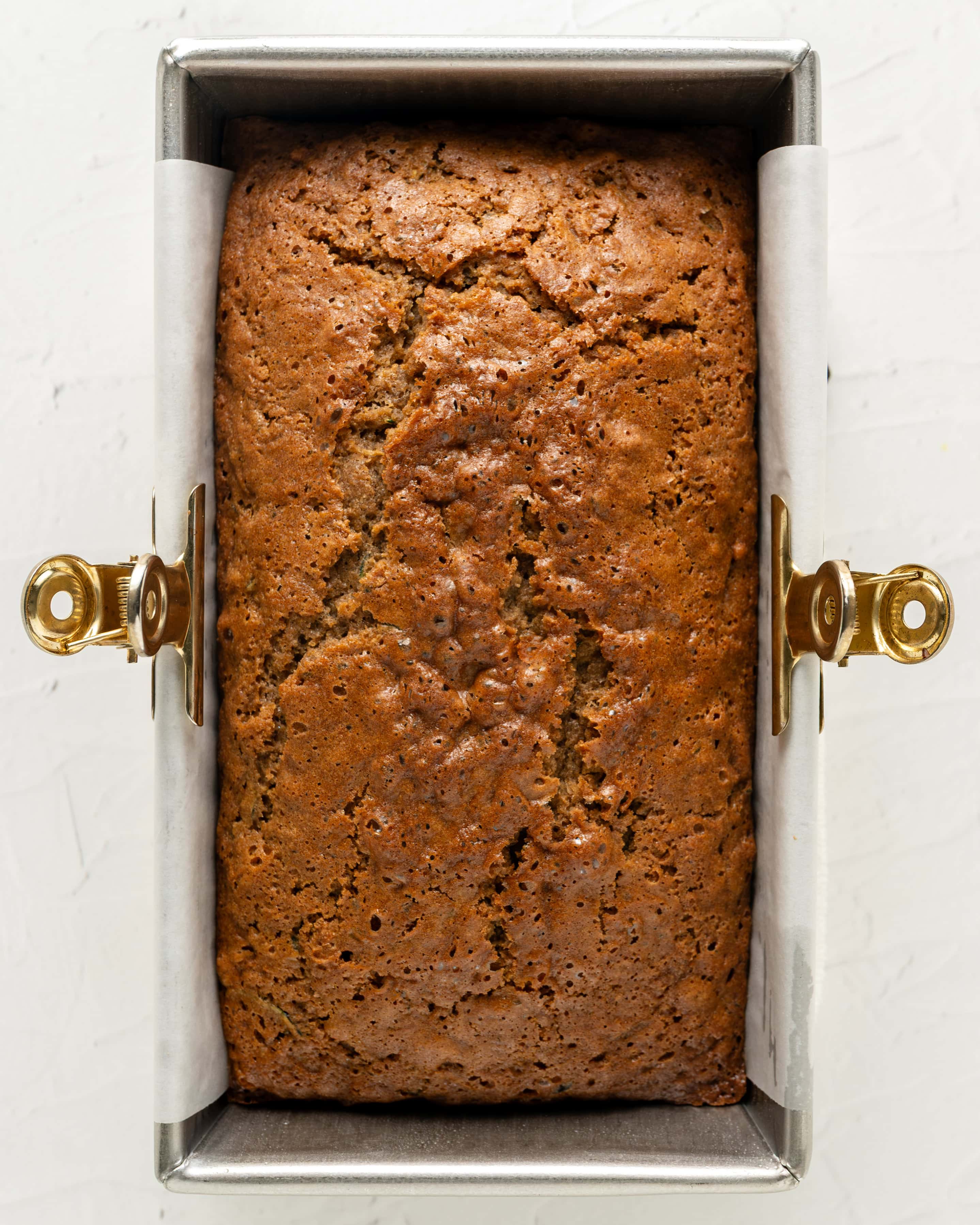 Overhead view of a freshly baked, golden-brown loaf of gluten-free zucchini bread cooling inside a parchment-lined metal loaf pan with gold clips.
