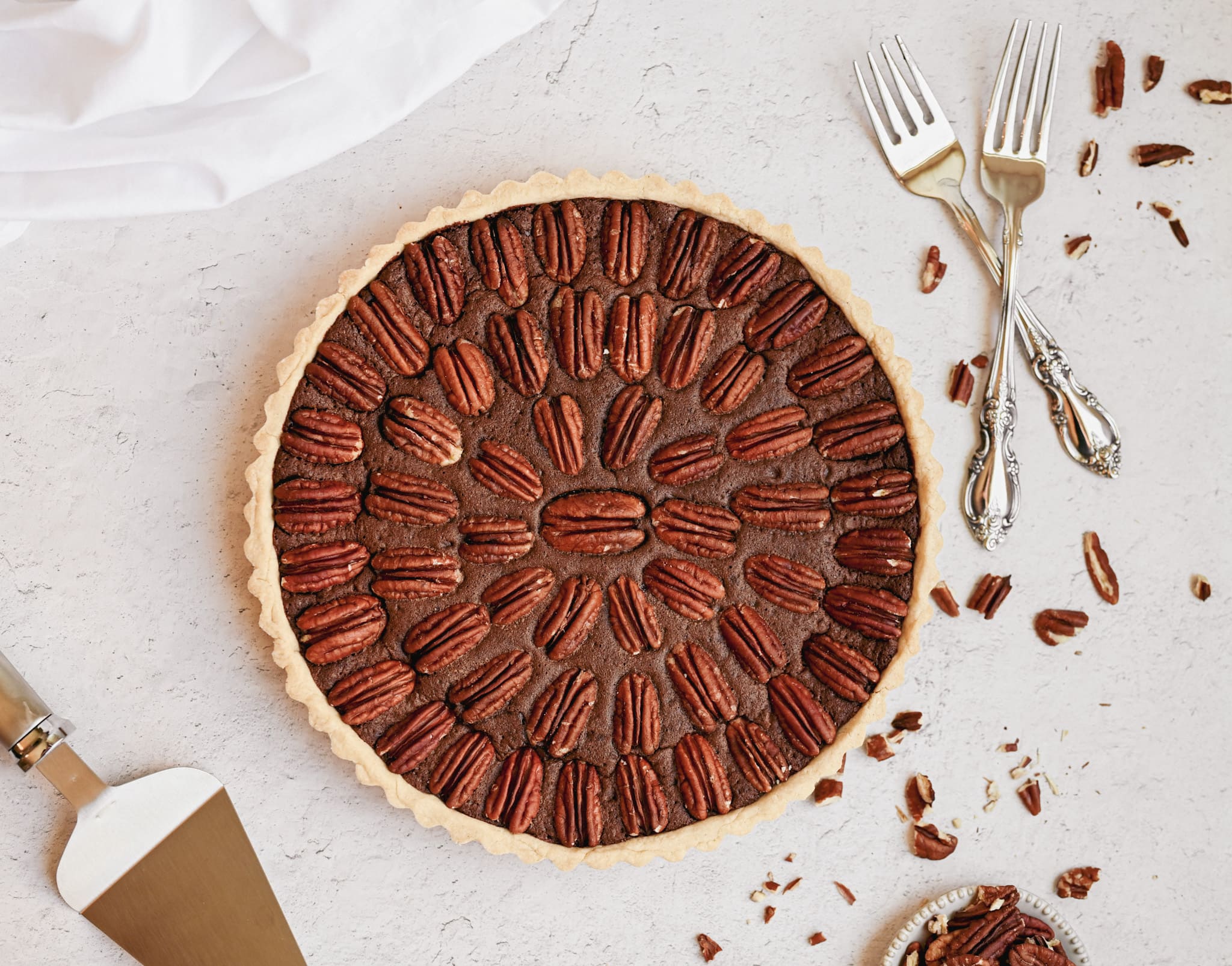 whole chocolate pecan tart shot from an aerial view, chopped pecans can be seen around the tart and white silverware