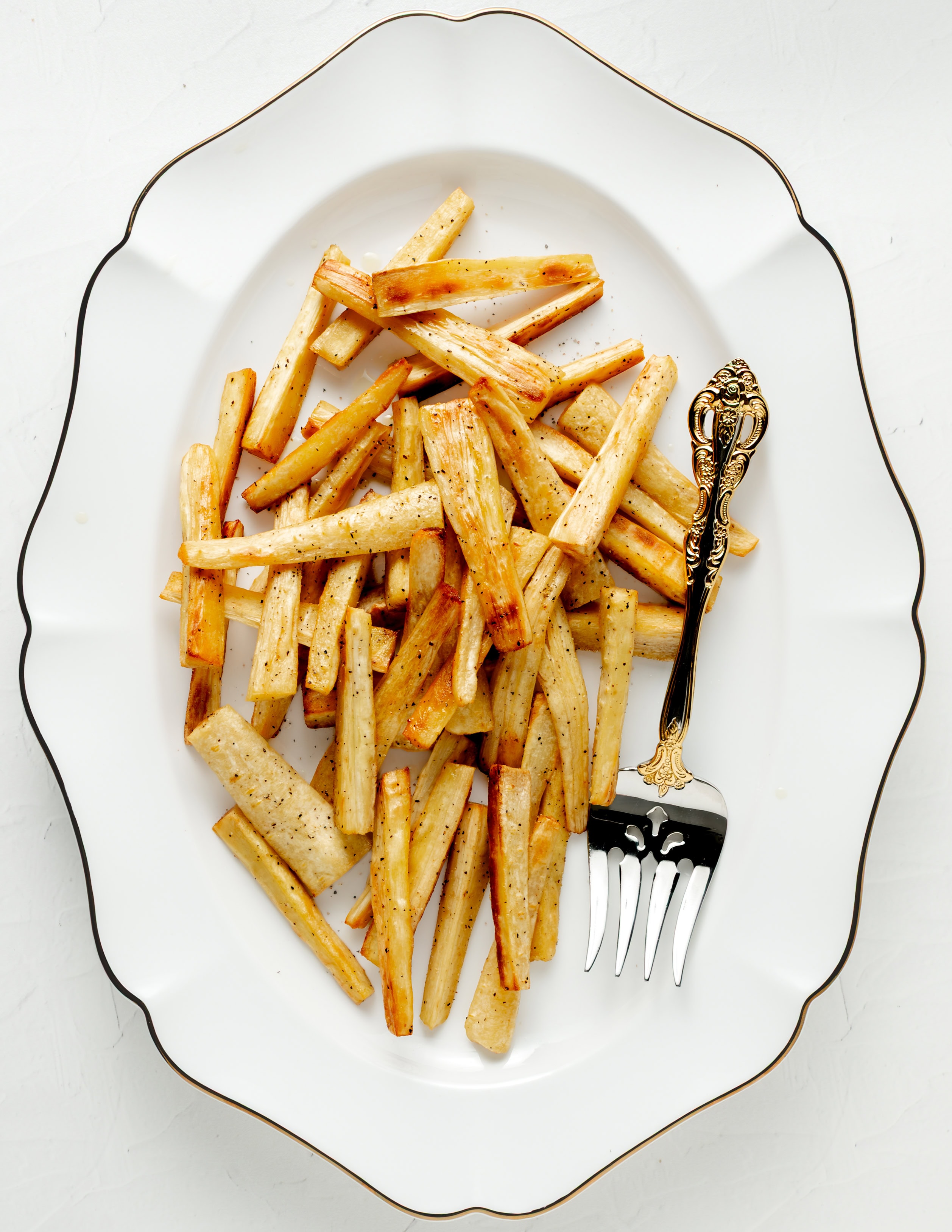 Roasted parsnips on a white platter with a gold and silver plated serving fork.