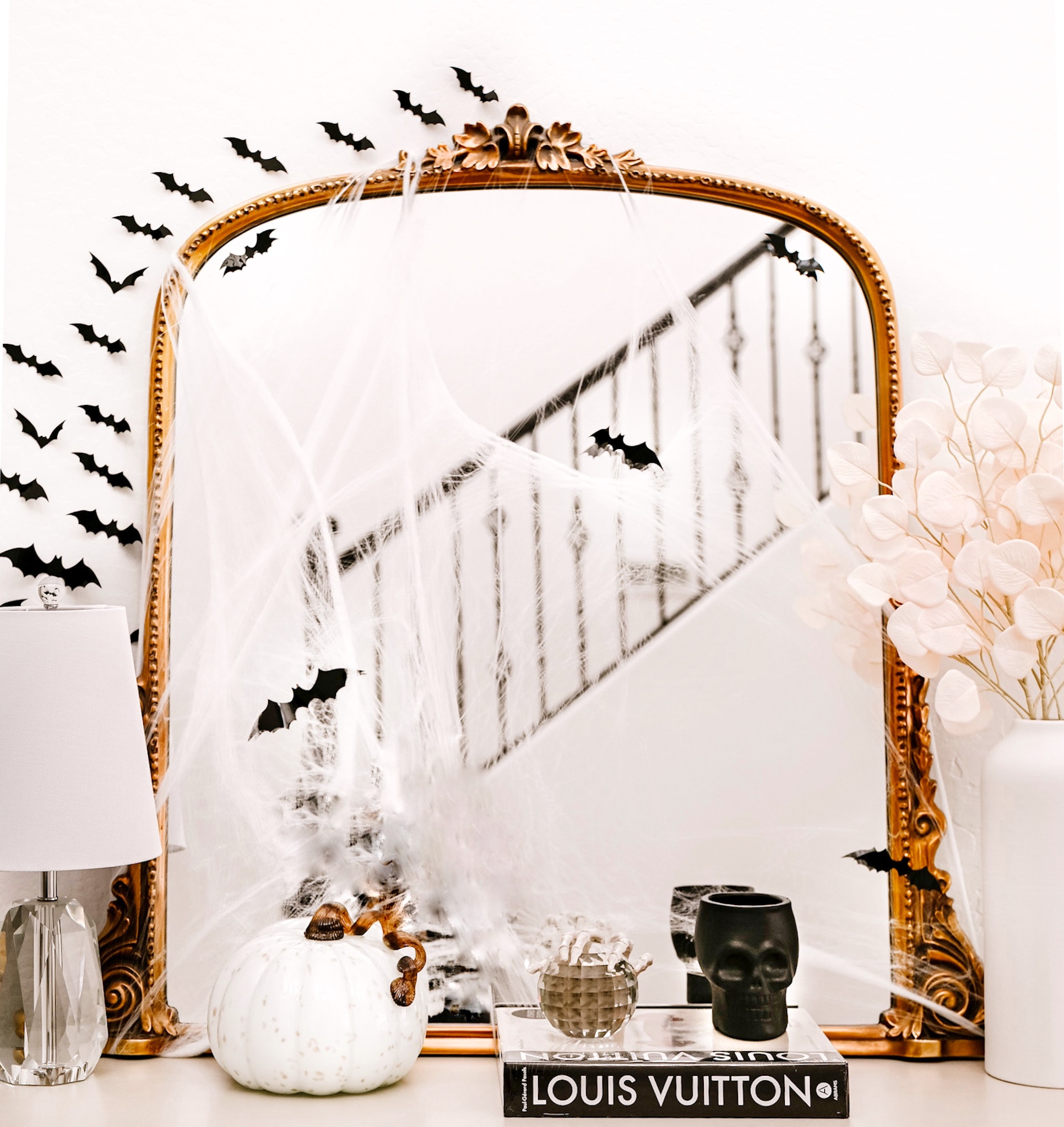 gold mirror with bats and cobwebs decorated on it. A white glass pumpkin sits on a dress next to a black skull, a crystal lamp, and white flowers in a vase