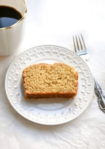 gluten free orange cardamom bread slice on a white decorative plate. A silver fork and a cup of coffee are next to the plate.
