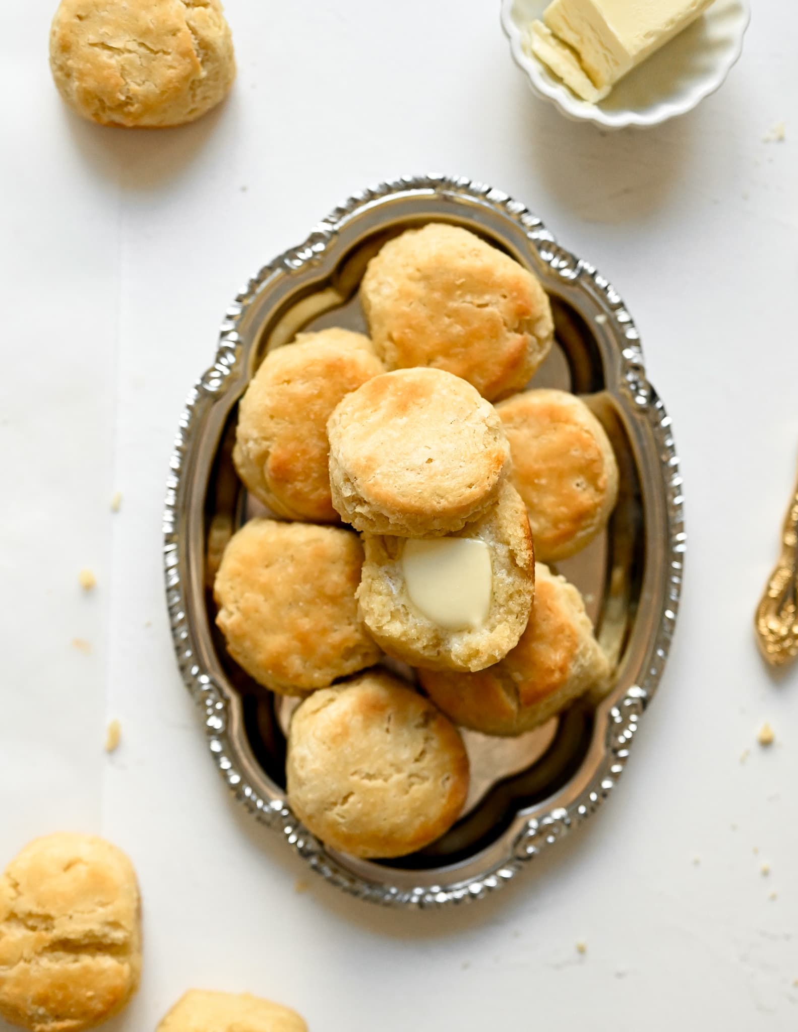 A pile of biscuits stacked on top of a silver ornate platter. The biscuit on the top is cut in half with melted butter. Surrounding the serving platter are several biscuits and butter in a serving dish.