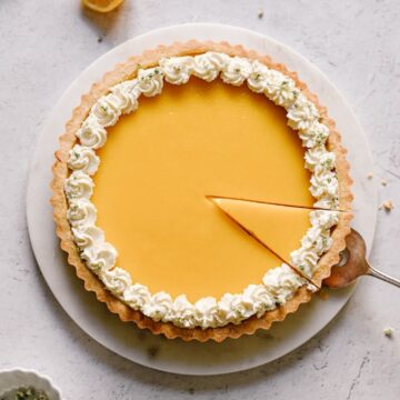 A whole gluten-free lemon tart with whipped cream around the edge. A single slice is being removed from the tart.