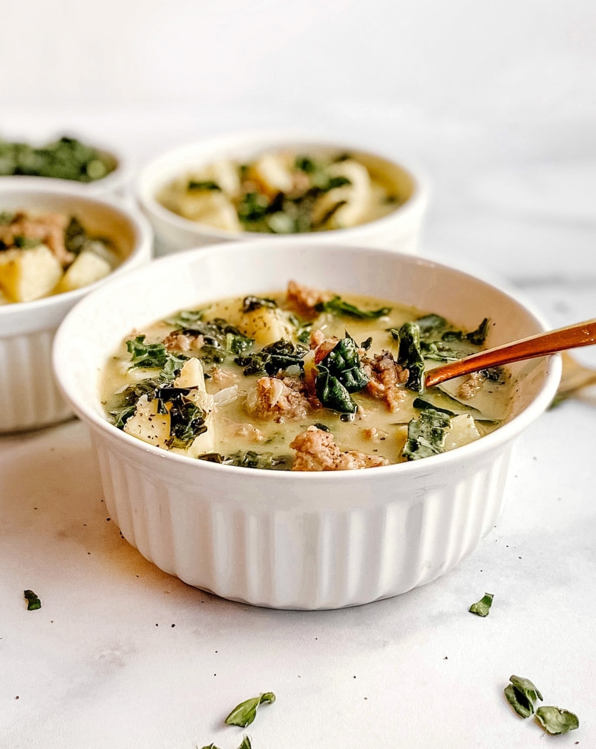 A bowl of gluten-free zuppa toscana soup with two additional bowls in the background. You can see chunks of meat, potatoes, and kale in the soup.