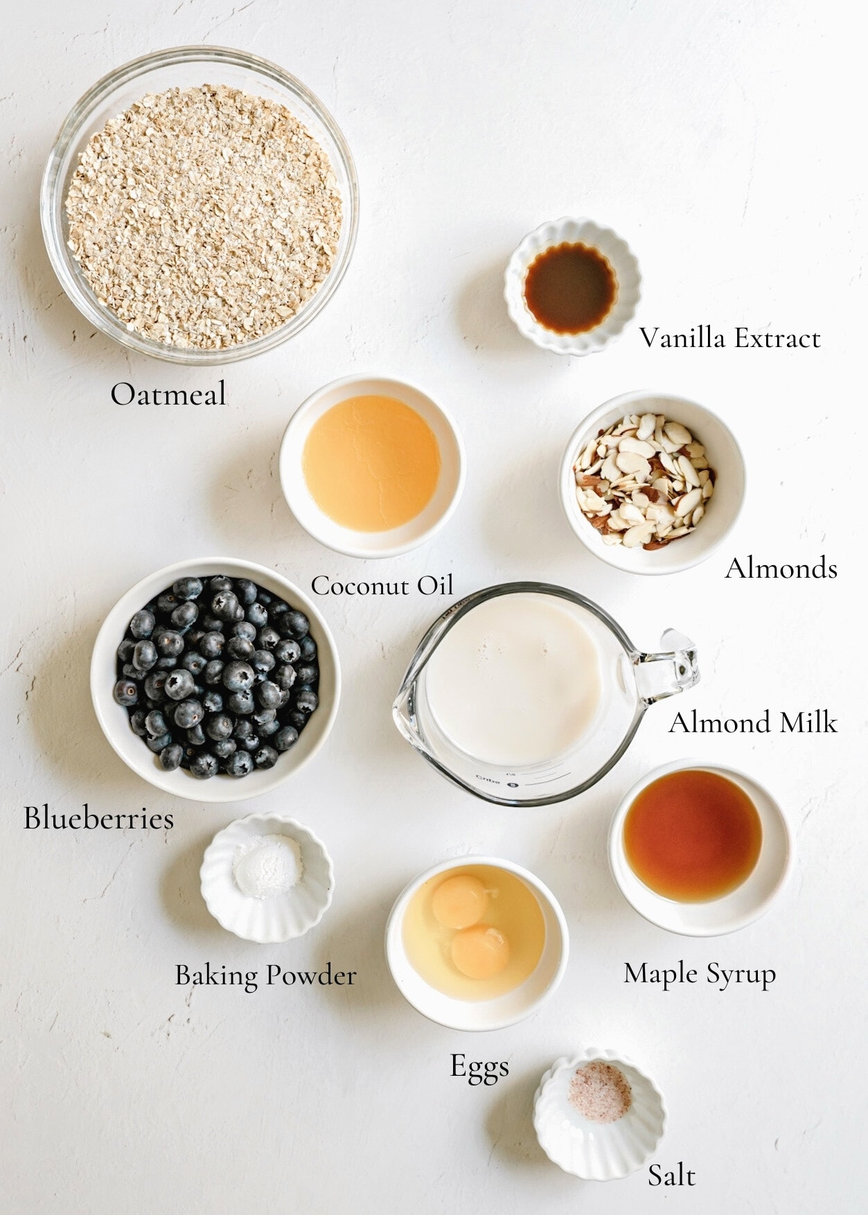 A photo of the various ingredients used to make the baked oatmeal. Ingredients include oatmeal, vanilla extract, coconut oil, almonds, almond milk, blueberries, baking powder, eggs, maple syrup, and salt.