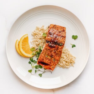 Salmon topped with an orange maple glaze on a bed of rice and micro greens. Two slices of oranges are used for decoration.