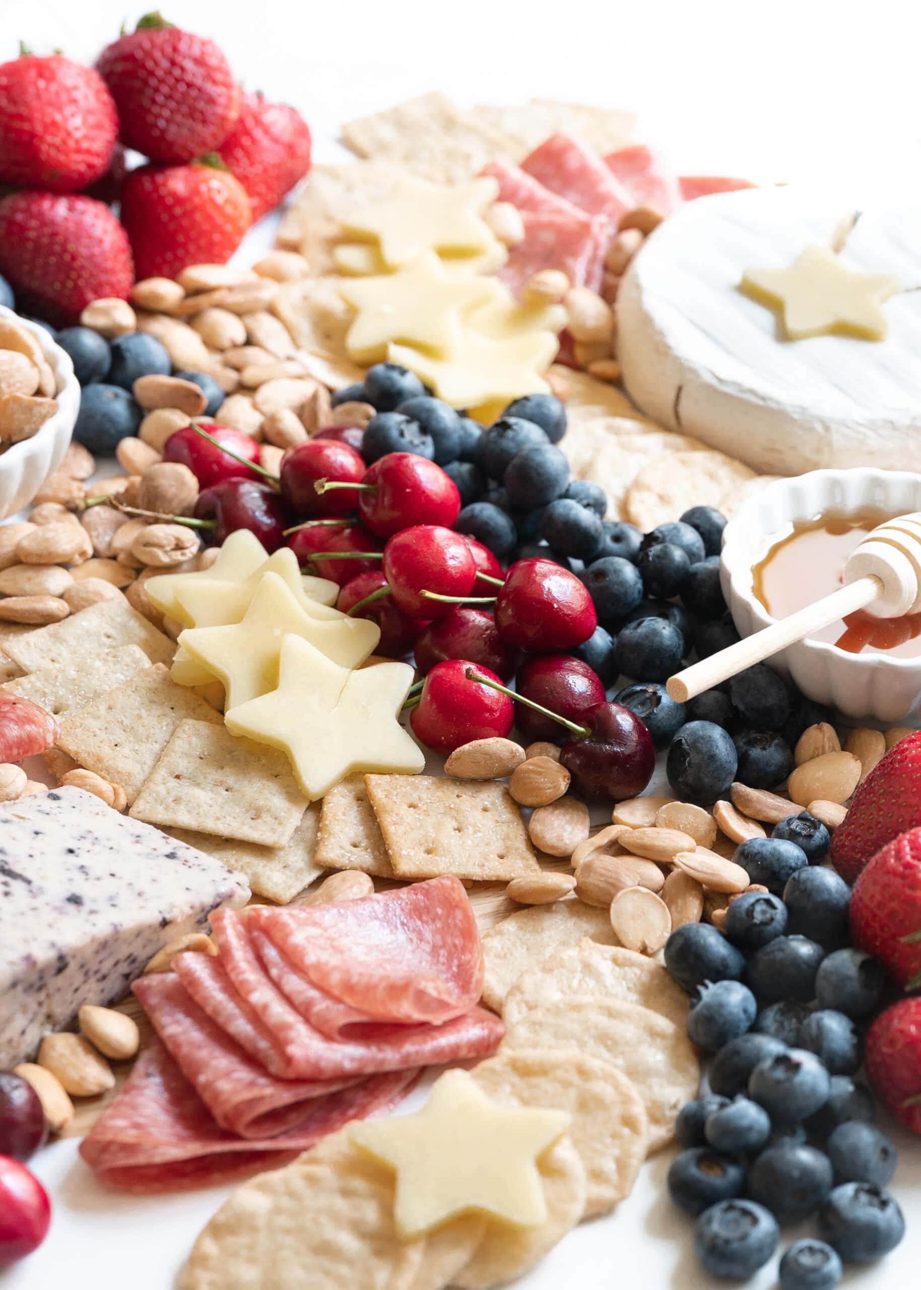 A photo of the patriotic charcuterie board from a front perspective. Gluten-free crackers, strawberries, blueberries, cherries, salami, white cheddar cheese, brie, blueberry cheese, almonds, and honey are on the board.