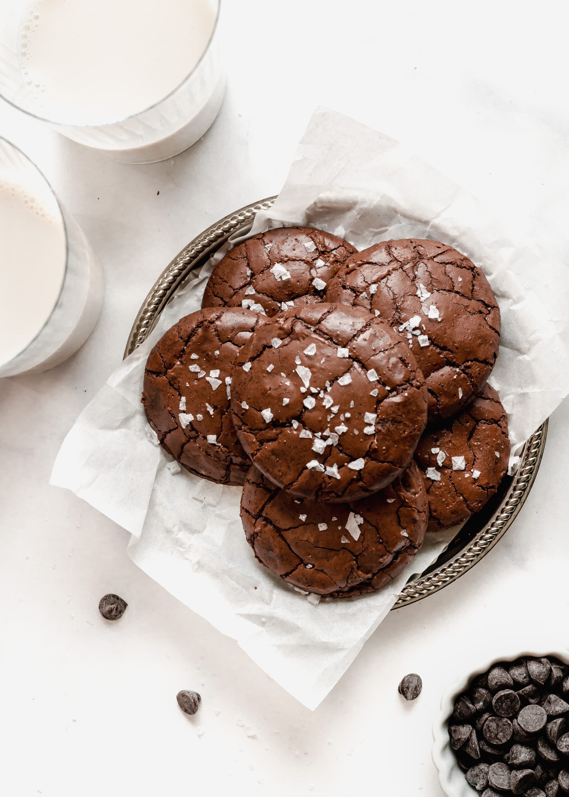 Six large chocolate brownie cookies on a silver platter with white parchment paper, next to two glasses of cold milk. Across from the silver tray in a small white bowl of dark chocolate chips.