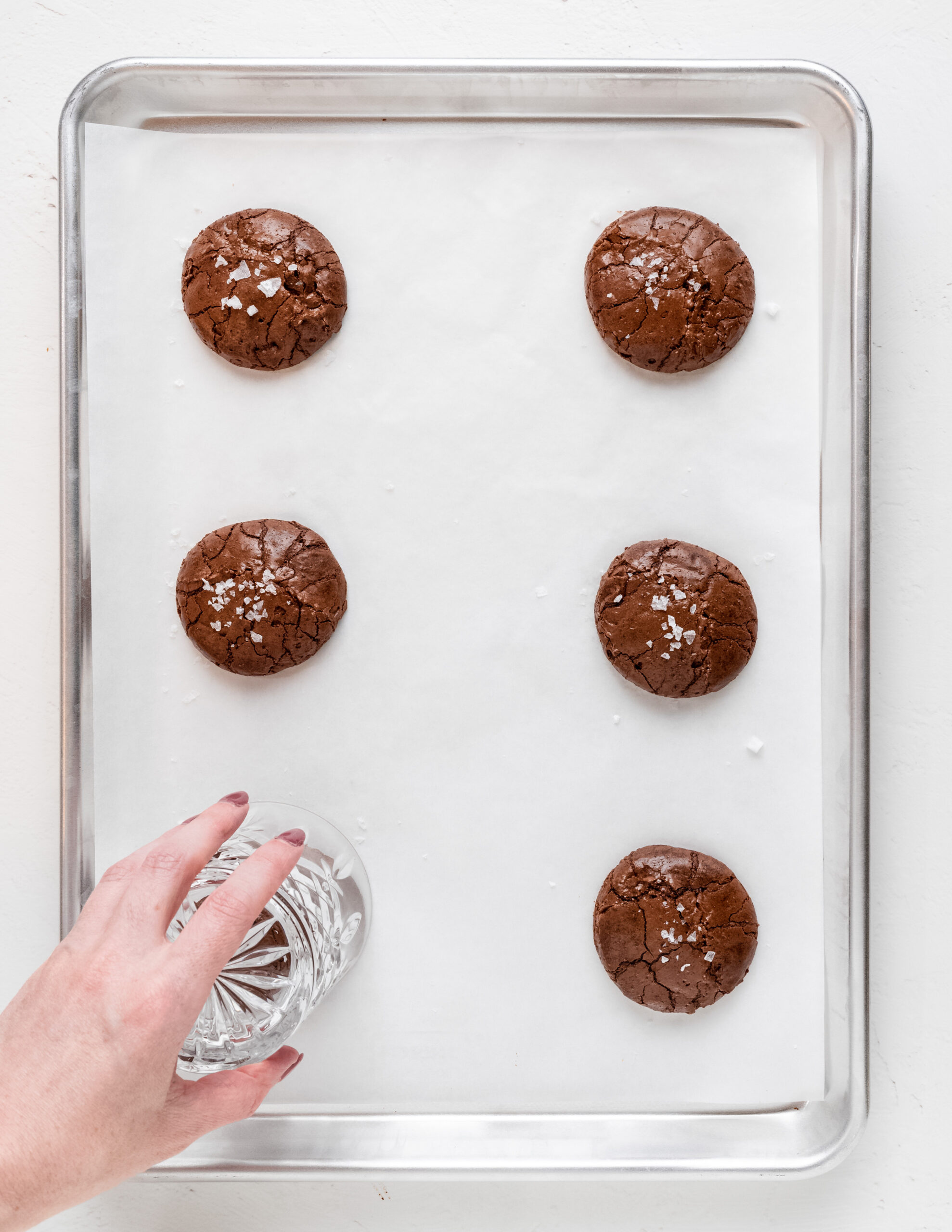 A finished product of 6 chocolate brownie cookies on a silver baking tray, lined with parchment paper. There's a visible hand with a glass over the top of one cookie, moving in a circular motion to create perfect roundness.