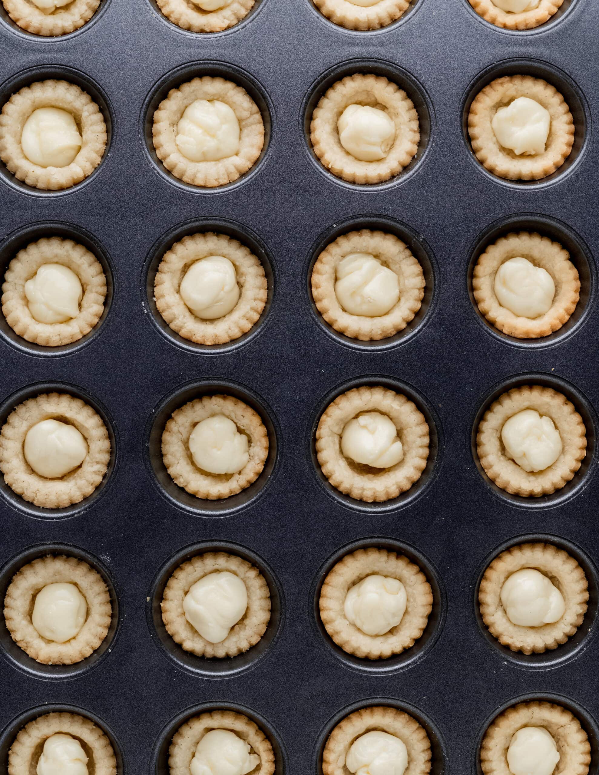 A silver mini muffin pan with baking tartlet shells and a chunk of brie placed in the middle of each tartlet.