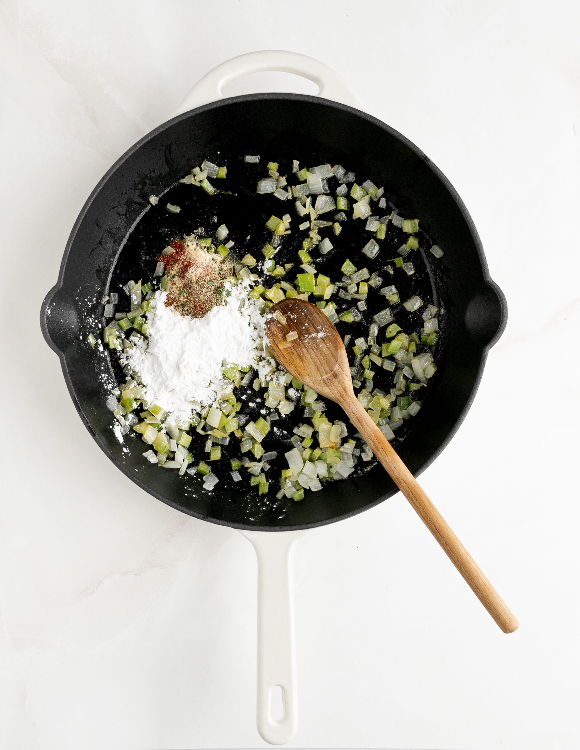 A large white skillet with onions, celery, tapioca starch, and seasonings, stirred with a wooden spoon.