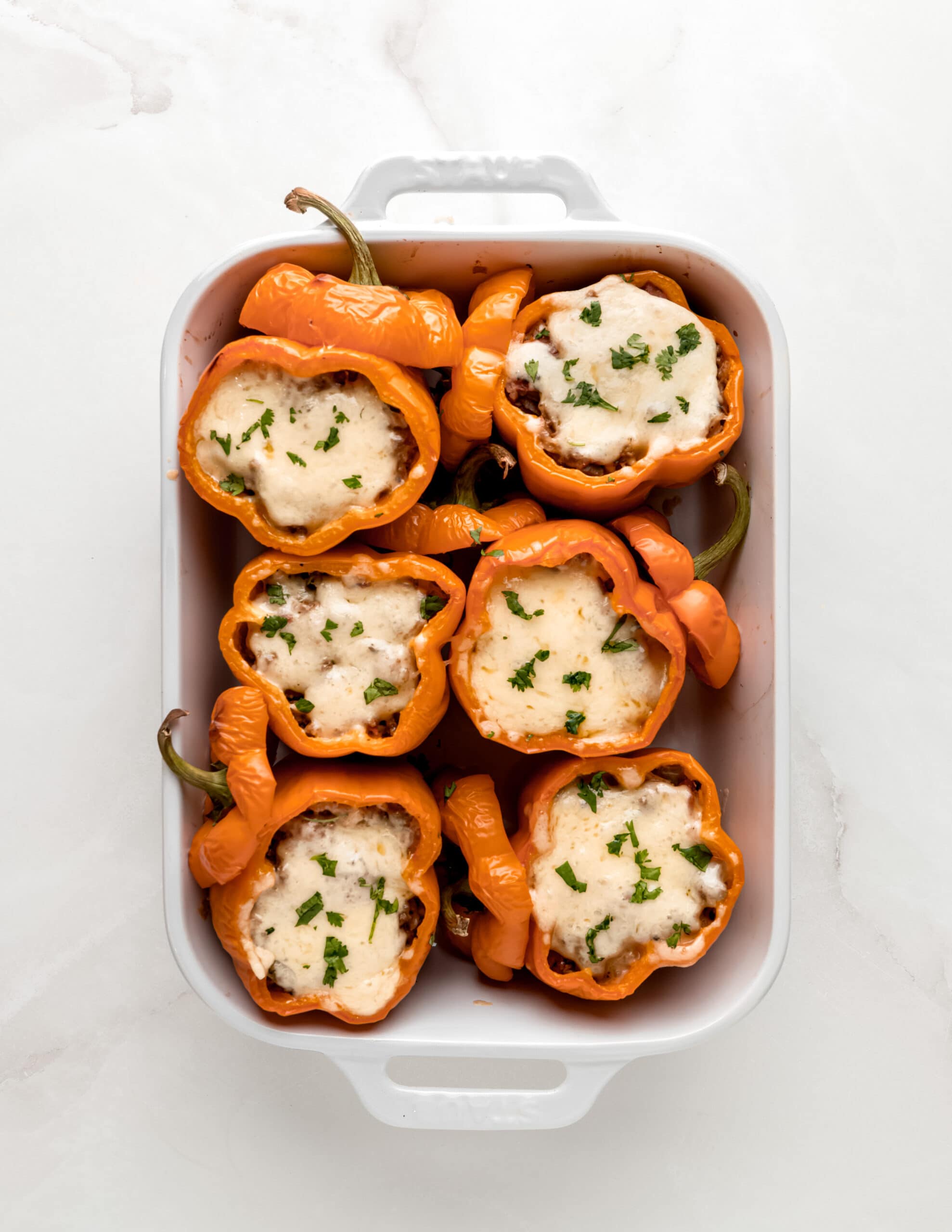 A large white ceramic baking dish with up-right orange bell peppers, fully baked, with cheese on top and cilantro to garnish.