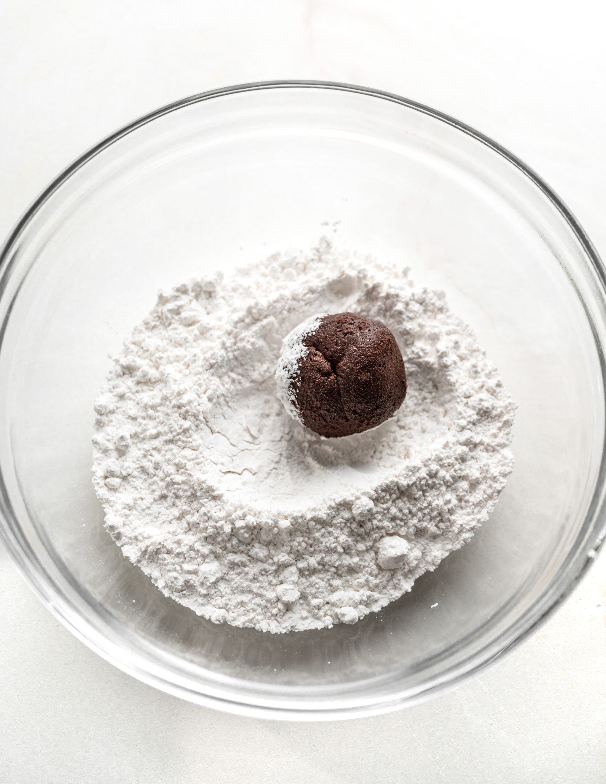 A large clear bowl with powdered sugar, and a ball of chocolate cookie dough being rolled in the middle of the powdered sugar. 