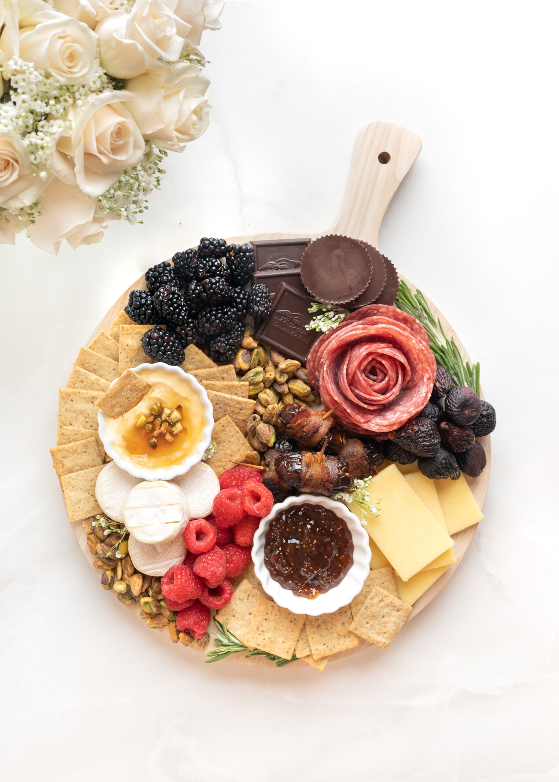 A Small charcuterie board with crackers, cheese, fruit, and chocolate. A vase with pink roses and babys breath are in the corner.