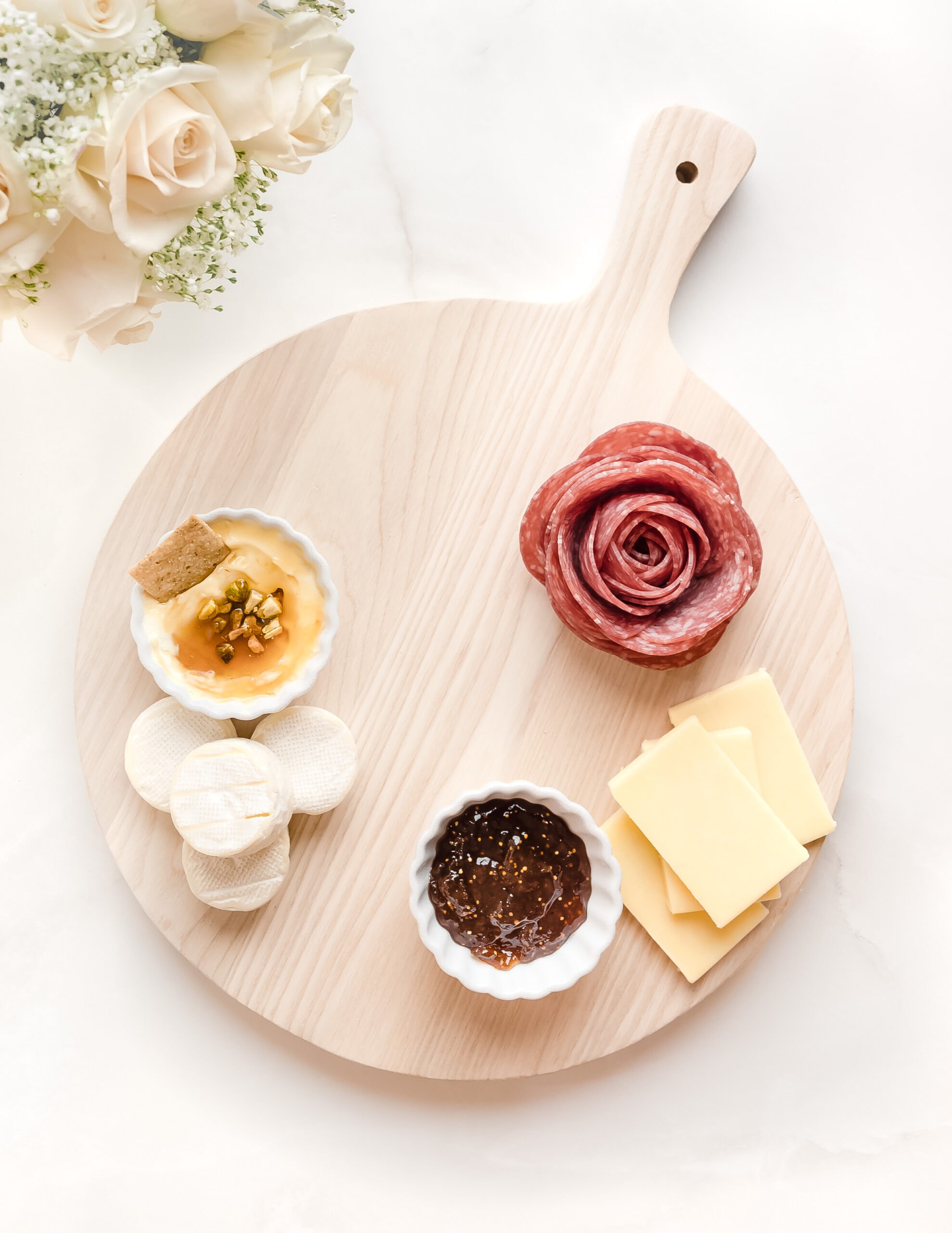 A small round wooden charcuterie board with marscapone cheese, fig jam, brie, cheddar, and a salami rose. Across from the board is a vase of pink roses. 