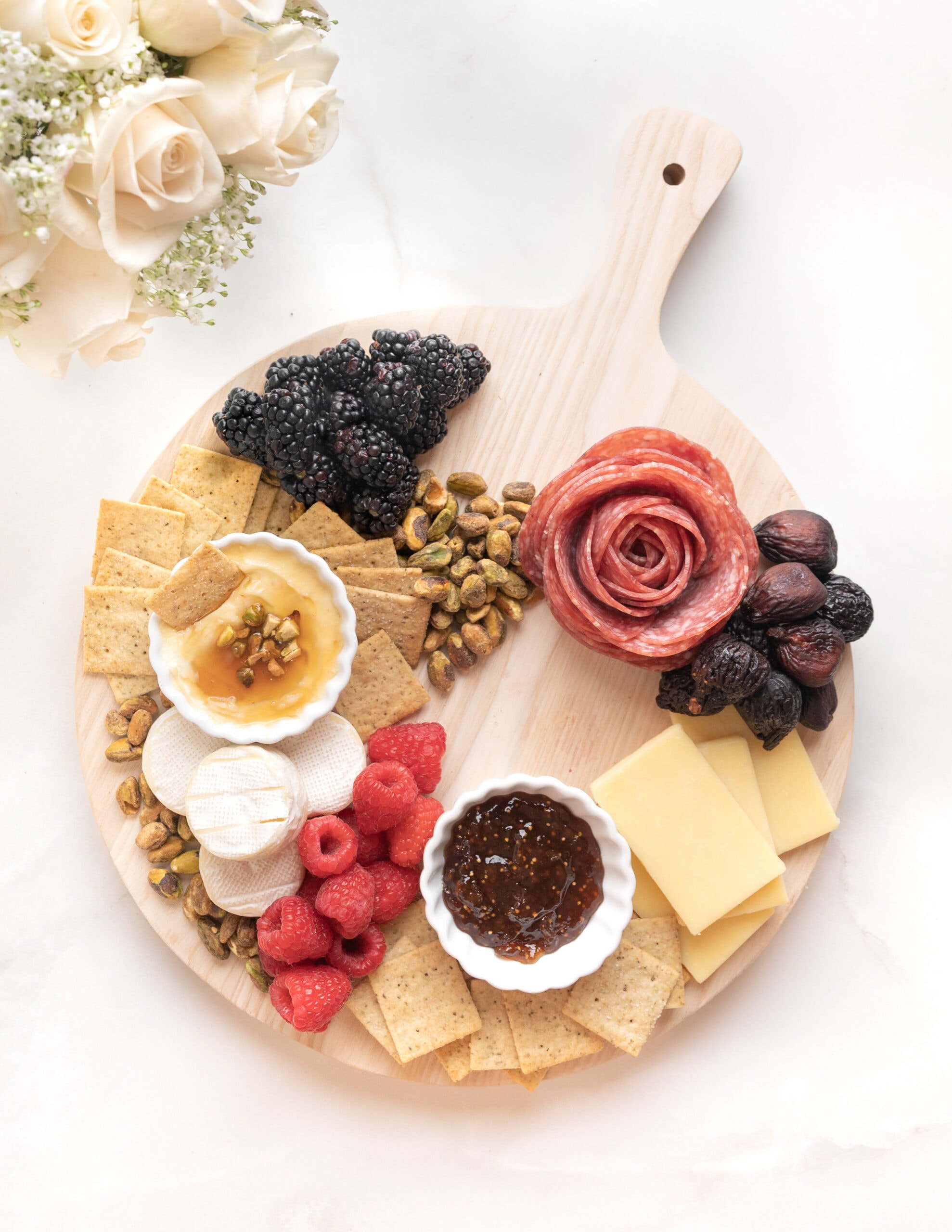 A small wooden round charcuterie board with marscapone cheese, fig jam, cheddar cheese, brie, crackers,fruit, and nuts. A vase of pink roses are across from the board. 