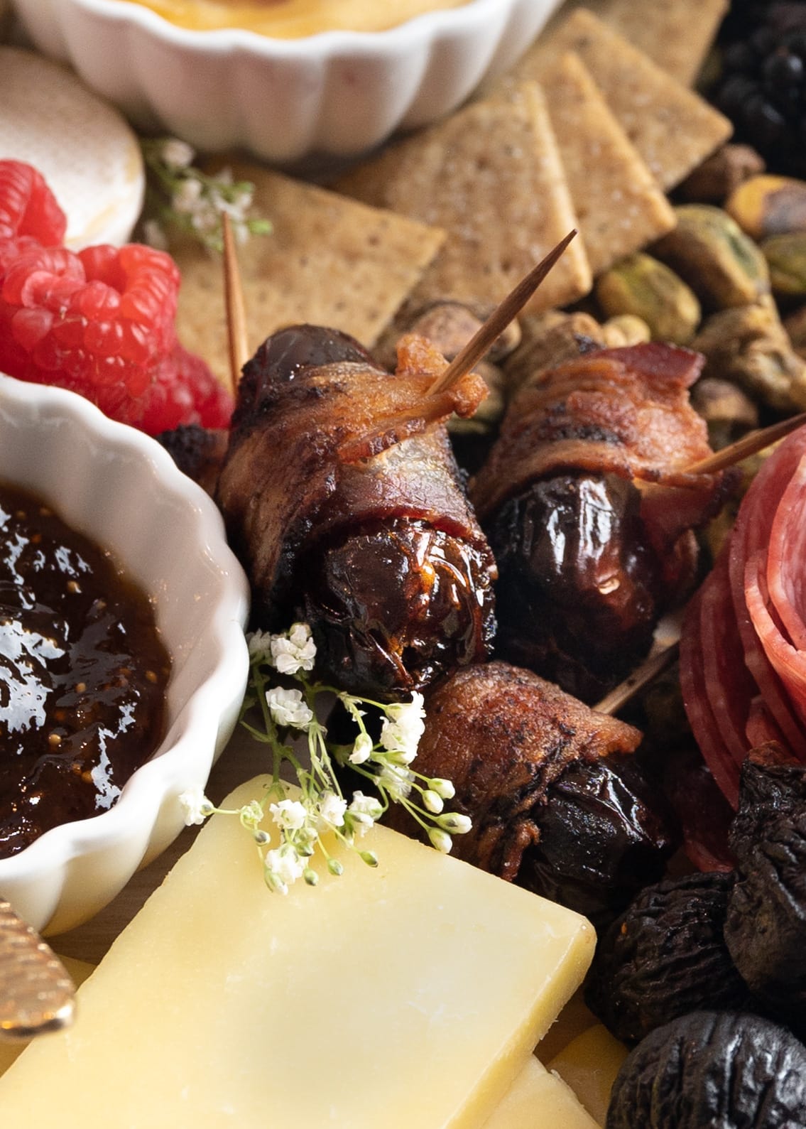 A close up picture of bacon wrapped dates with cheddar cheese, babys breath flowers, fig jam, pistachios, crackers, and dried figs.