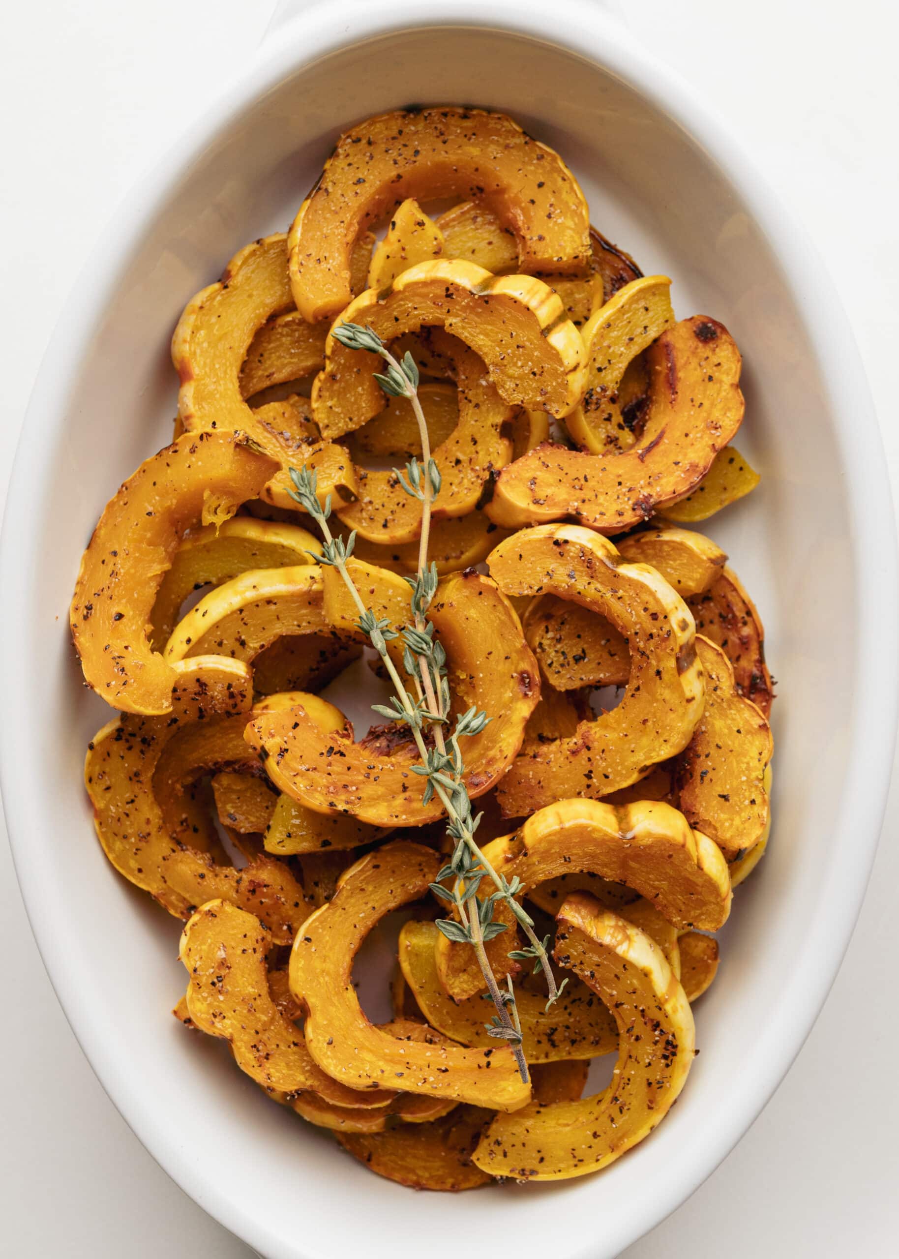 A large white oval dish with rings of delicata squash sprinkled with salt, pepper, and springs of fresh thyme in the center.