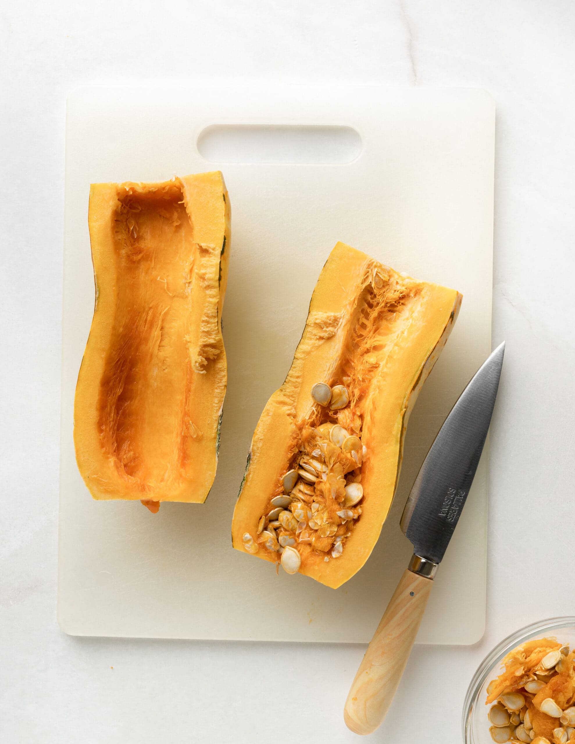 A white cutting board with a sliced open delicata squash, partially scopped seeds with a knife in the corner.