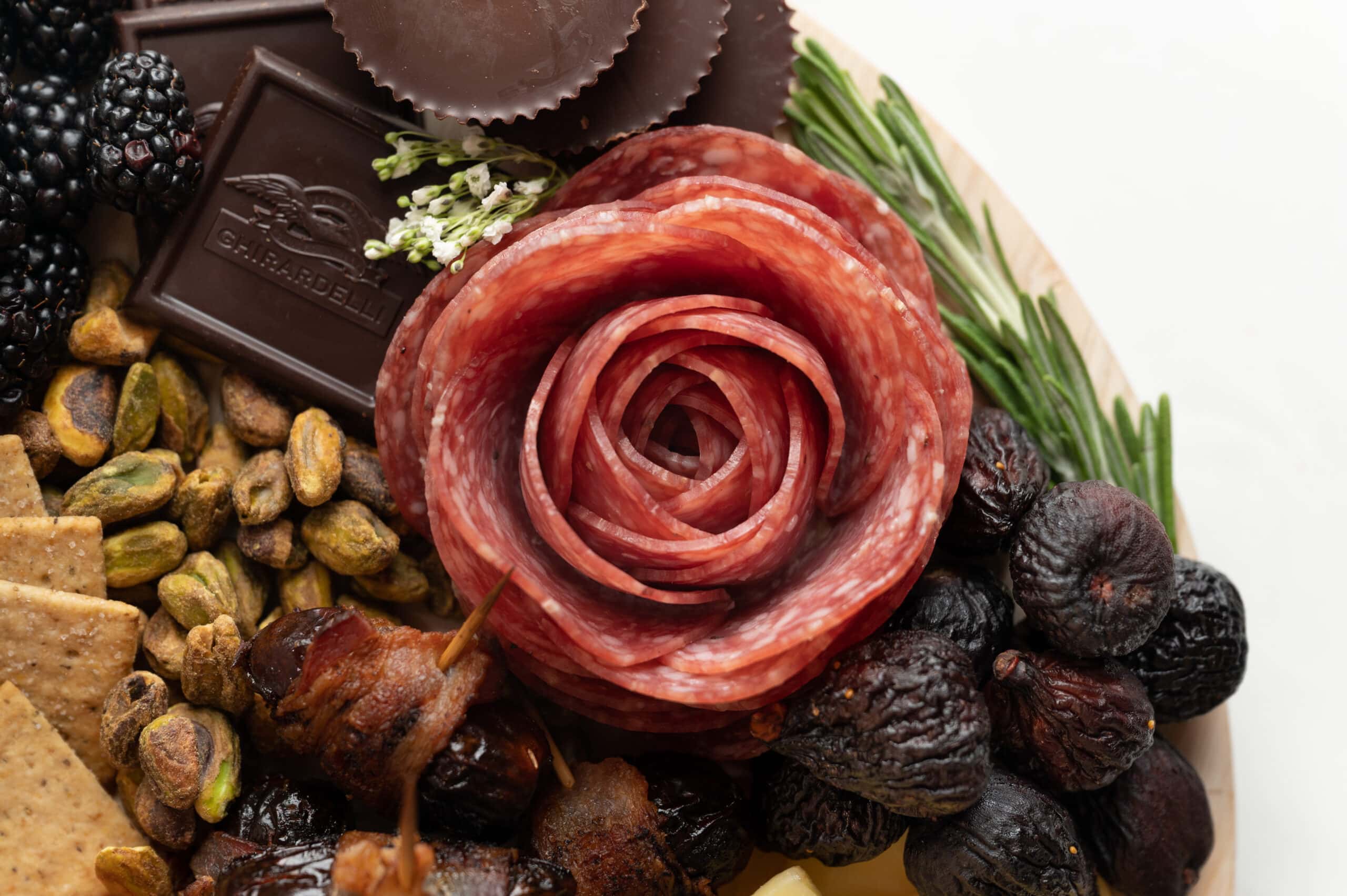 A picture of a salami rose on a charcuterie board with bacon wrapped dates, figs, rosemary, pistachios, crackers, and chocolate. 