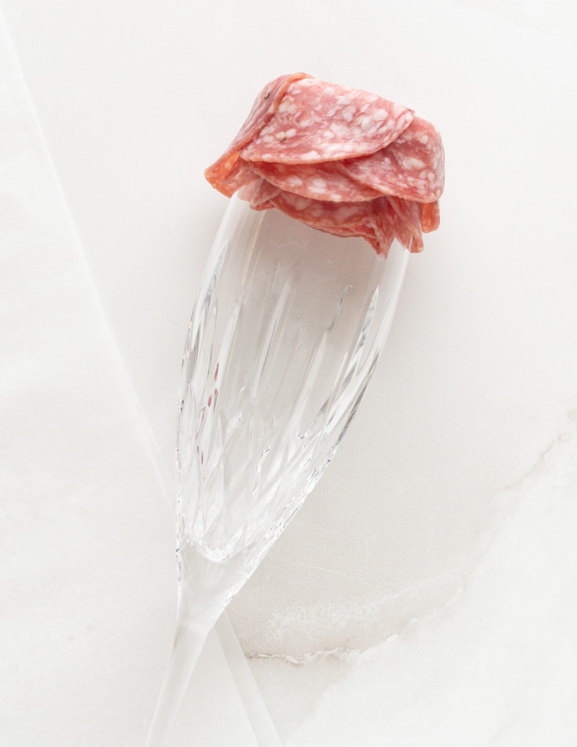 A champagne flute with several layers  of salami on the rim of the glass, layering on top of one another.