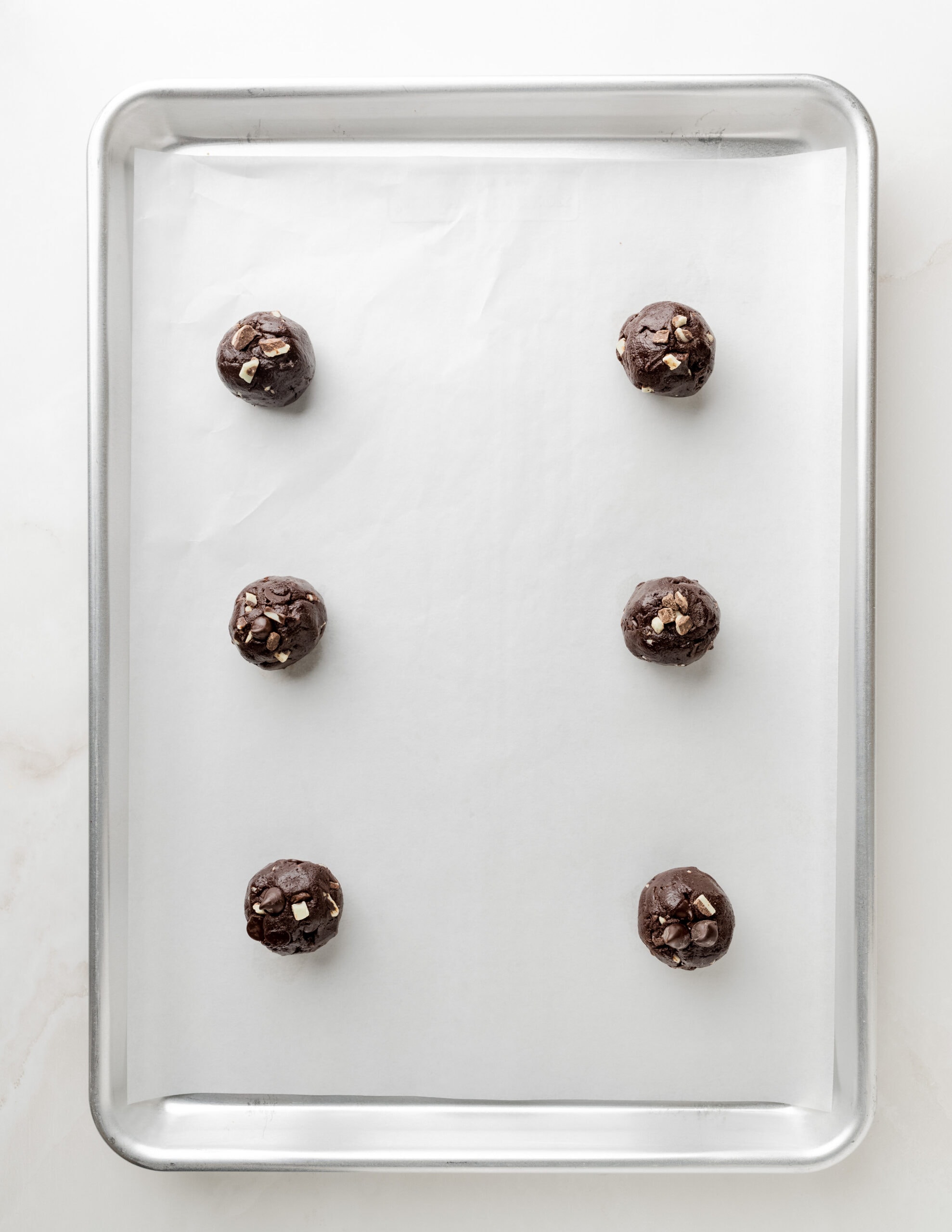 A large silver baking tray lined with parchment paper and six round balls of mint chocolate cookie dough.