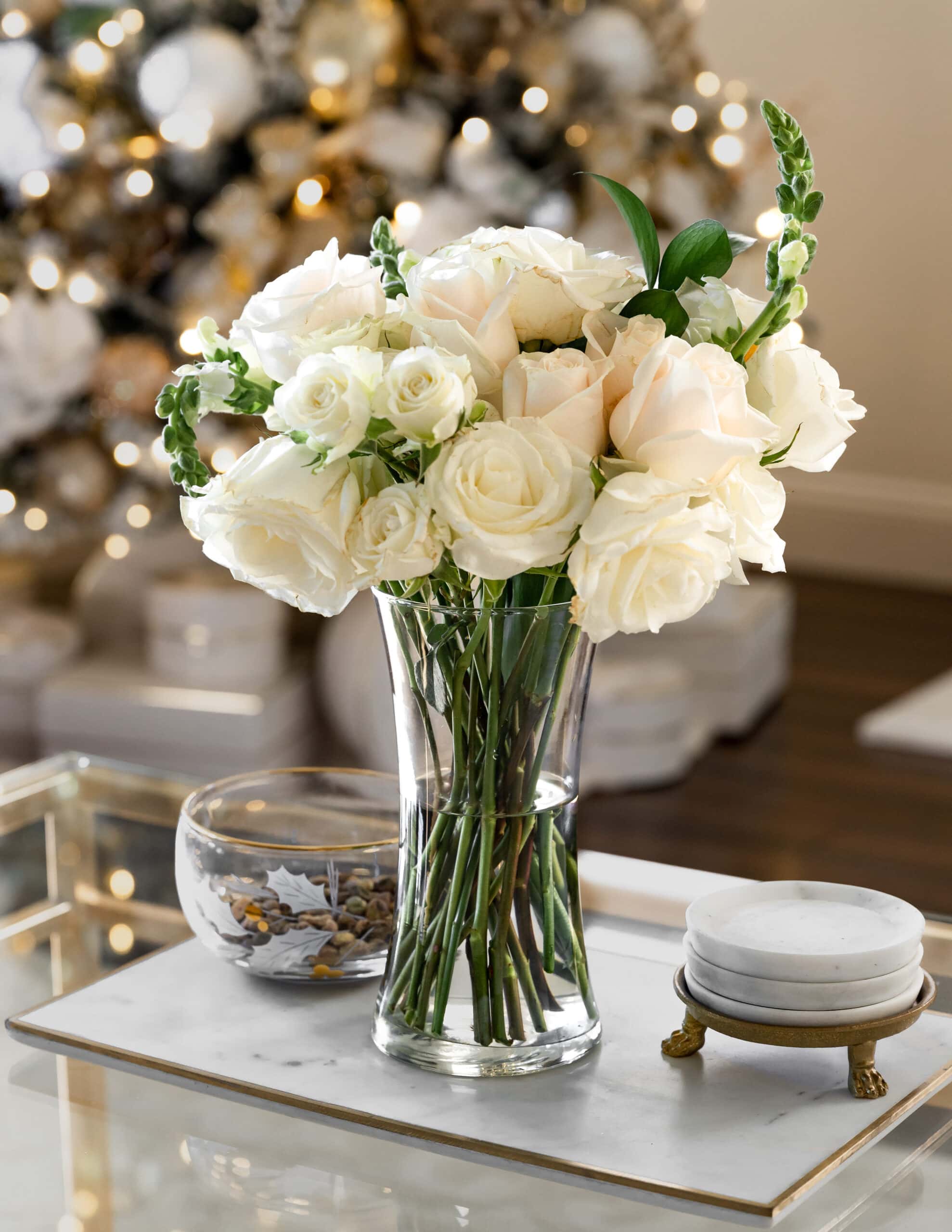 A bouquet of white and blush roses with green stems, sitting on a marble stand, white a gold claw footed coaster holder and a glass bowl off to the side.