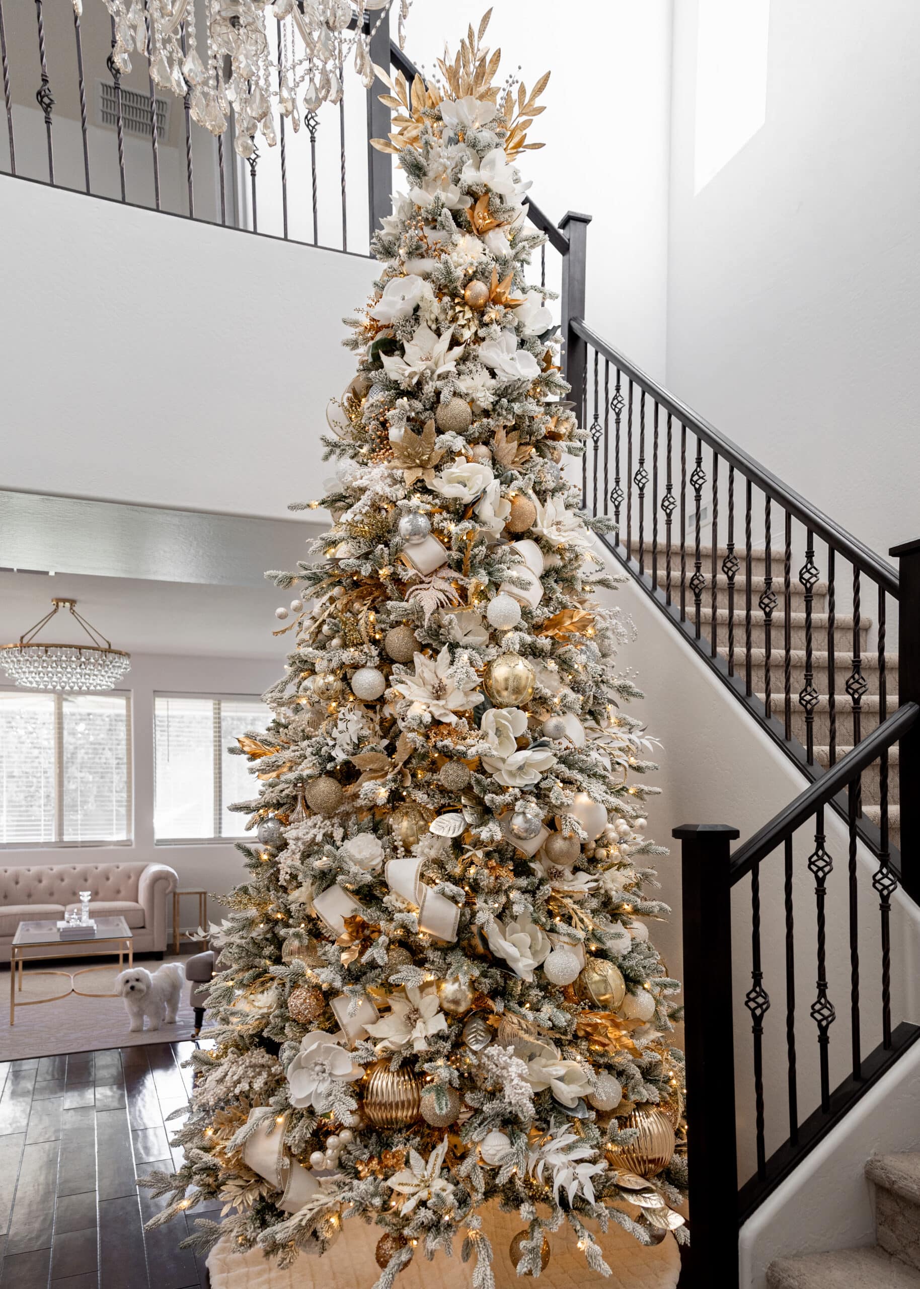A large 12 foot white and gold christmas tree with white and gold ornaments, white decorative ribbon, and a white dog in the background. 