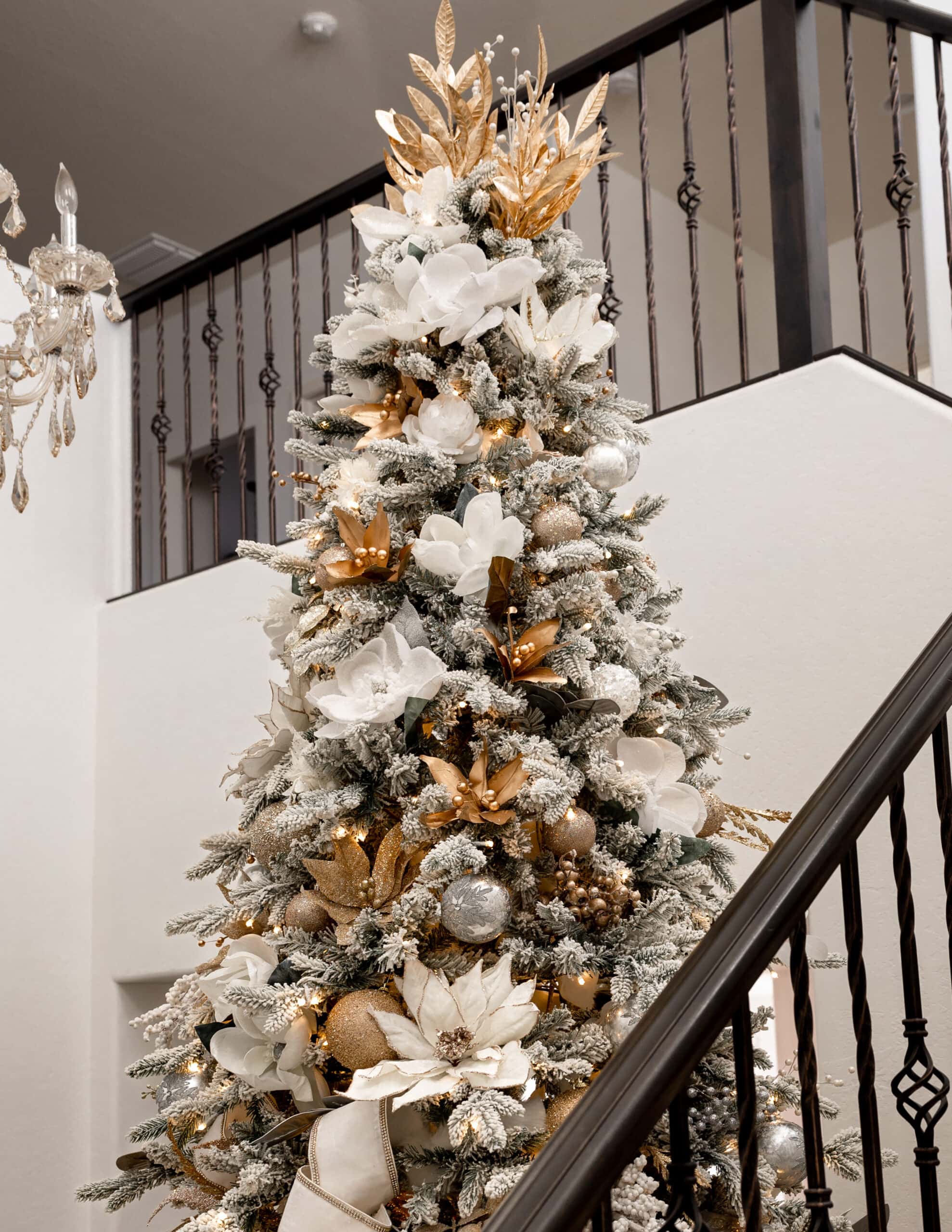 A close up of a 12 foot christmas tree with white and gold florals throughout, and a silver chandelier next to the top of the tree.