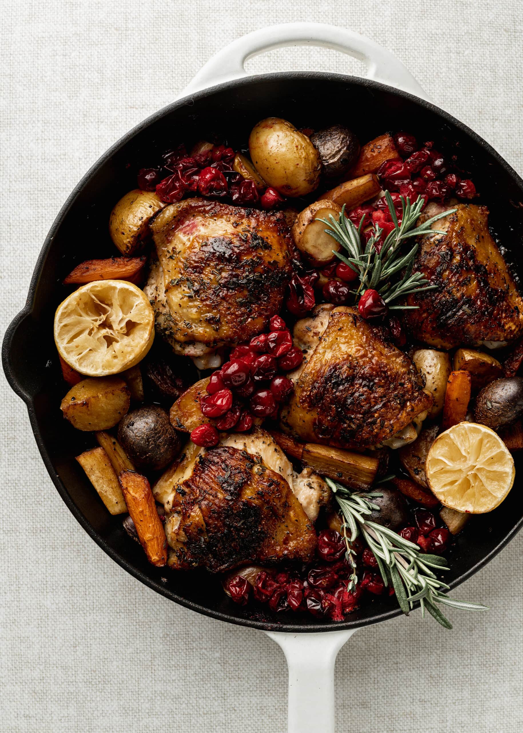 A large white skillet with with potatoes, carrots, and parsnsips with chicken thighs, rosemary, and cranberries.