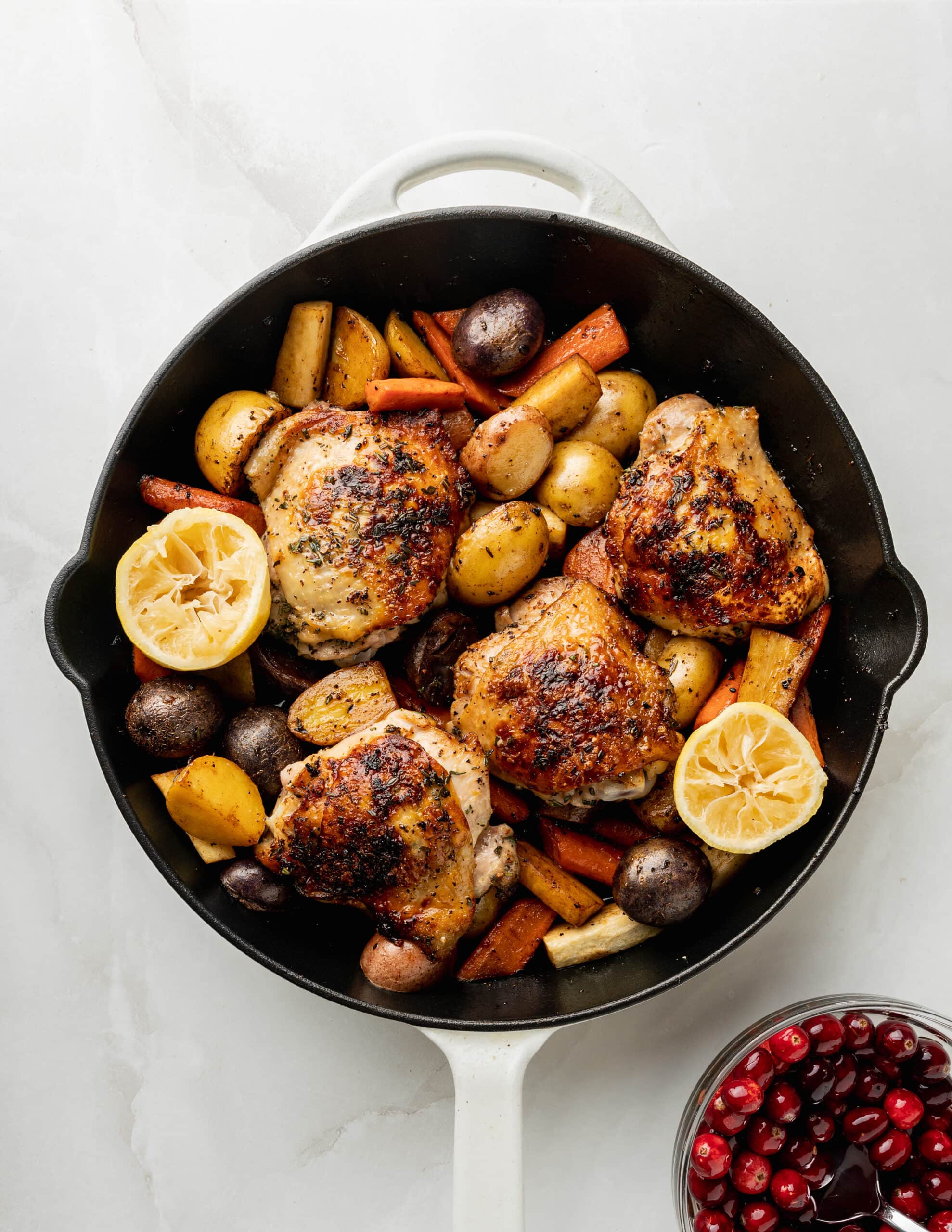 A large white skillet with potatoes, carrots, and parsnips, with chicken thighs seared on top. 