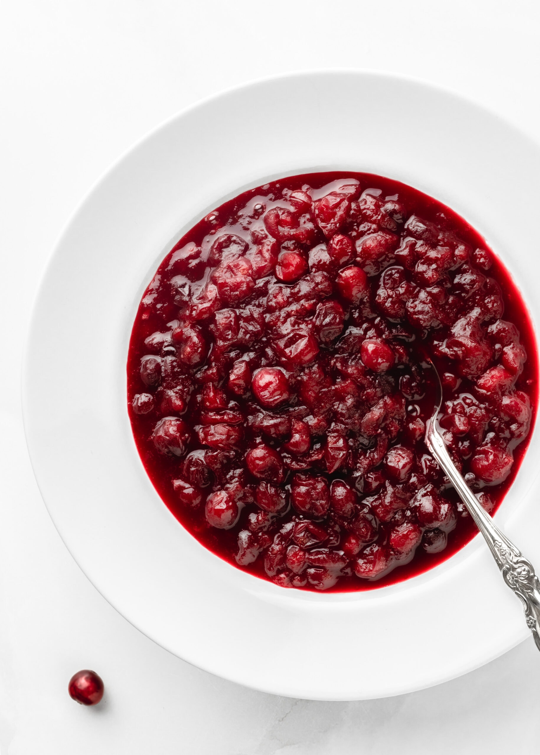 A large white bowl filled with vibrant red cranberry sauce, served with an antique silver spoon placed inside the bowl.