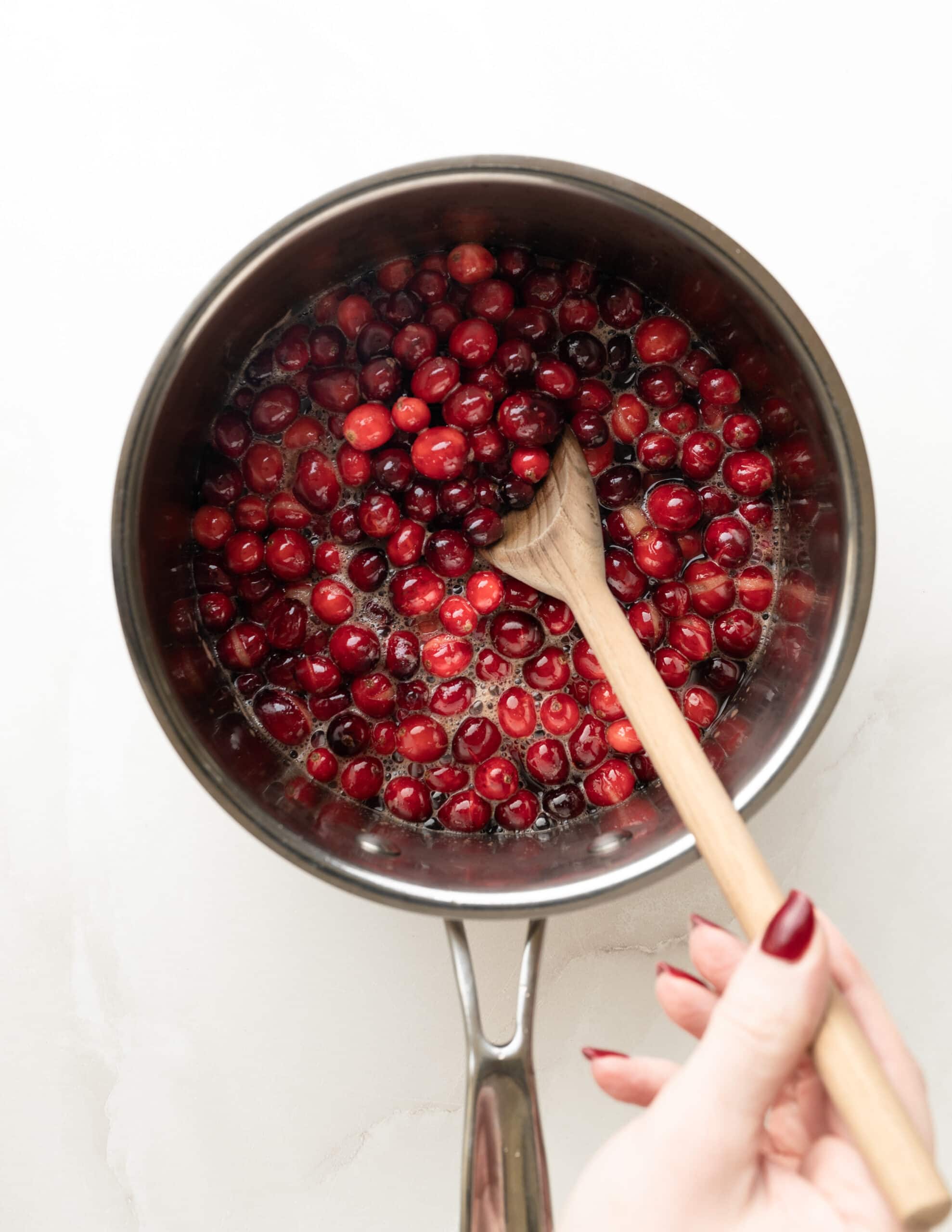 A stainless steel pot with orange juice, red wine, maple syrup, and cranberries, being stirred with a wooden spoon.