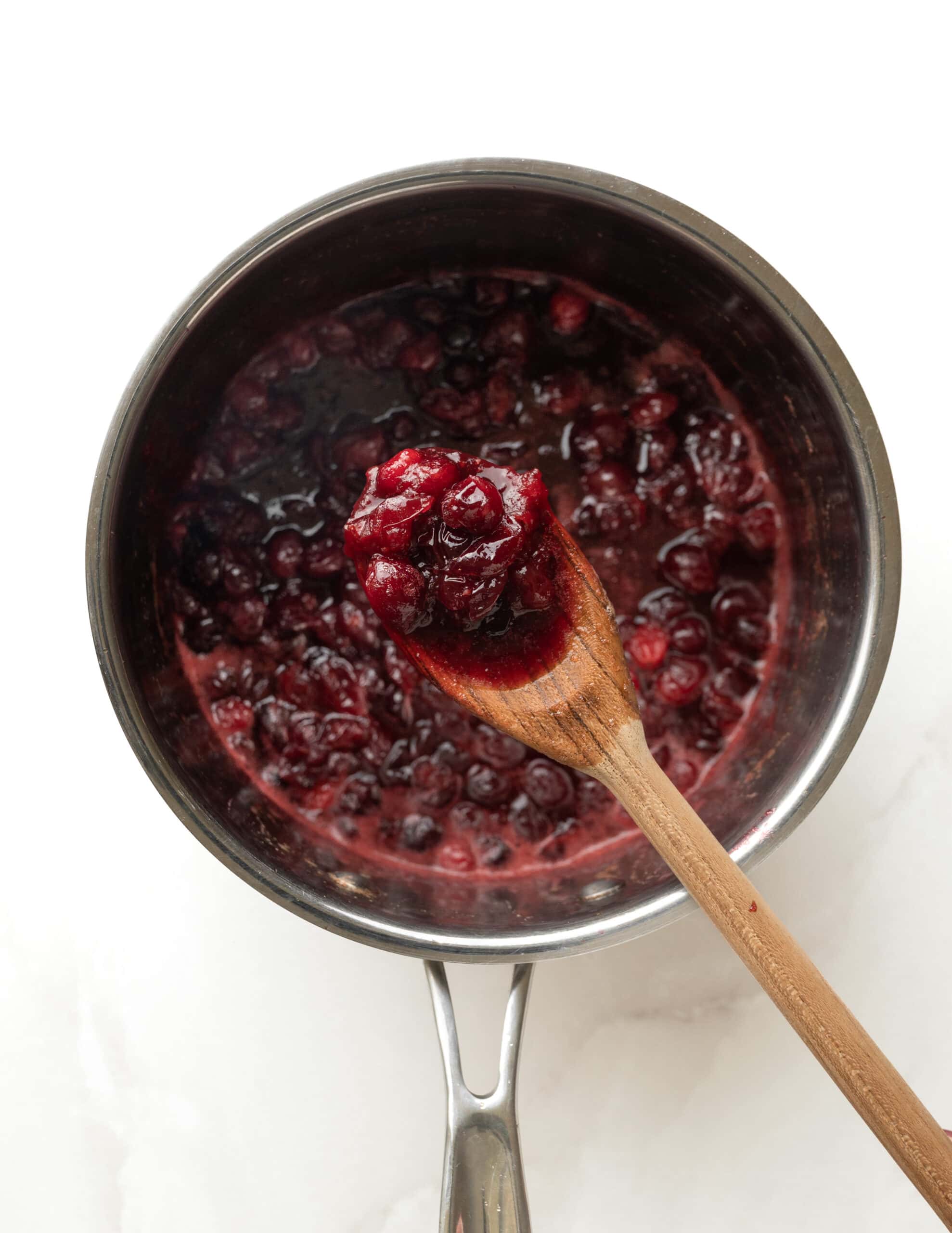 A stainless steel pot with cranberry sauce being held up by a wooden spoon.