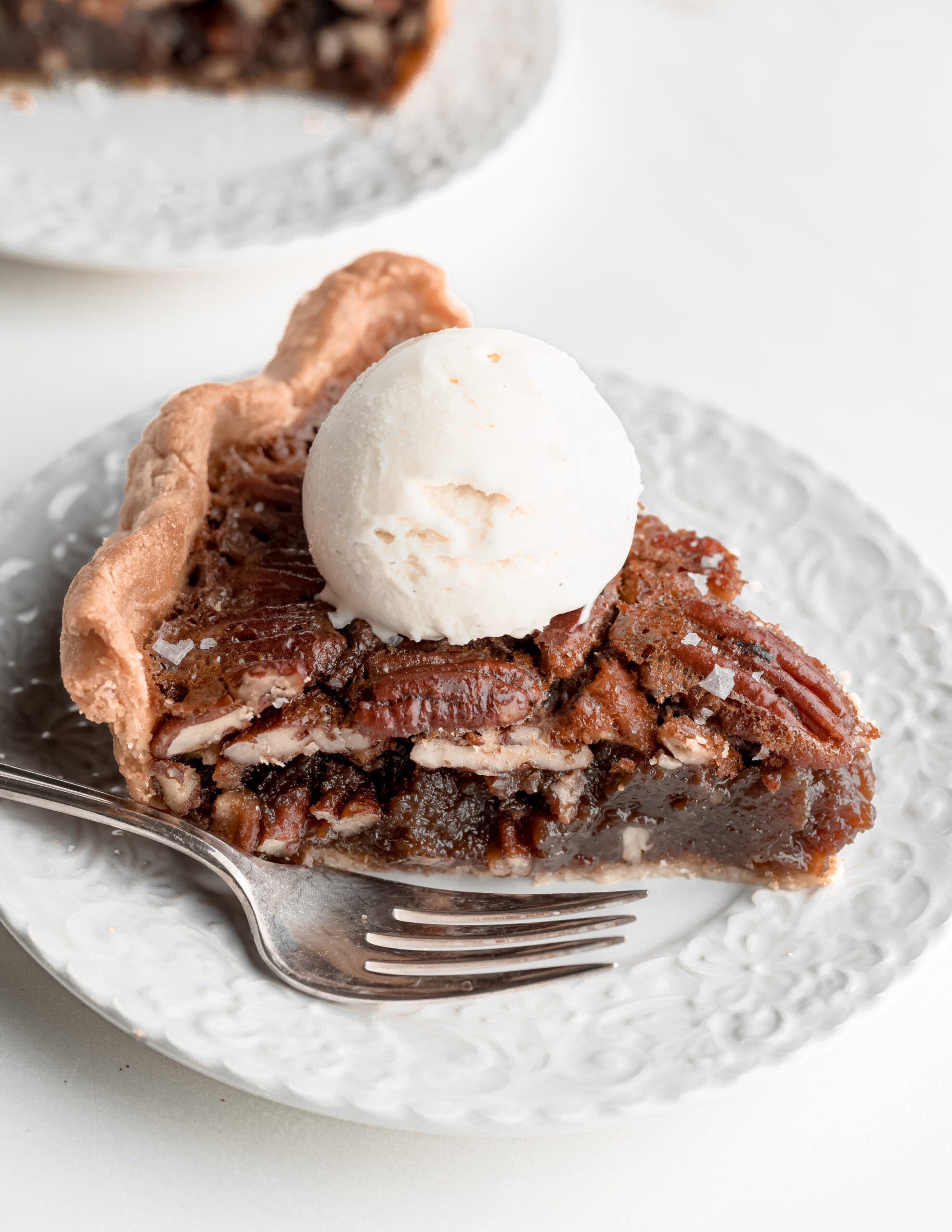 A large picture of a piece of pecan pie on a ceramaic white plate with vanilla ice cream on top.